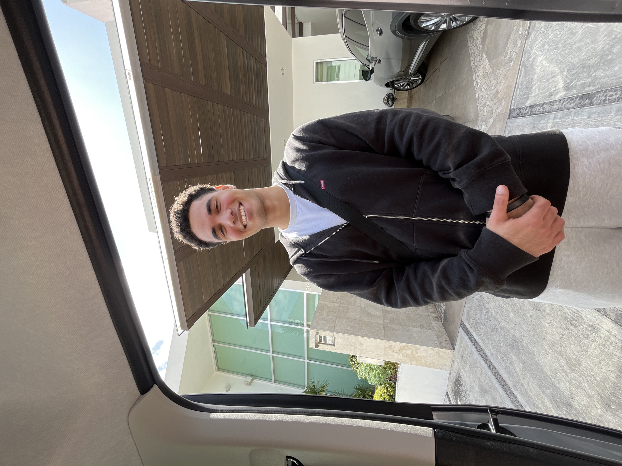 A young man stands smiling in front of a modern-style house in San Bernardino Tlaxcalancingo, Mexico. He is wearing a black zip-up hoodie over a white t-shirt and grey sweatpants. A black strap is visible across his chest, possibly from a bag. The house has a large window pane facade, and a dark wooden garage door. A silver car is parked in the driveway. The photo is taken from inside a car.