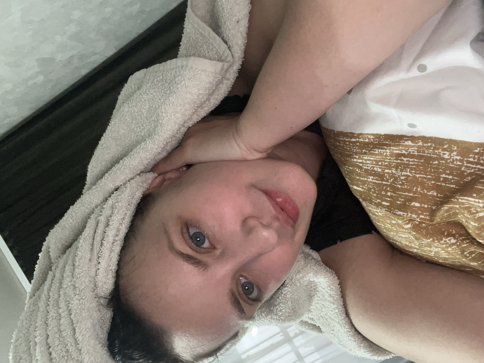 The media file is a close-up, vertically oriented selfie of a person wrapped in a white towel. The person's face is the central focus, with their head tilted and eyes looking towards the camera. They have light skin, blue eyes, and pinkish lips. A white towel is draped over their head and shoulders. Part of their arm is visible, resting on their chest. Behind them, a patterned fabric, possibly a pillowcase or blanket, with a design of white dots and horizontal brown lines is visible. The background includes a dark, textured surface and a lighter, possibly tiled surface. The lighting suggests an indoor setting, possibly a bathroom or bedroom. The person's expression appears relaxed or contemplative. There is no visible text in the image. The location context of Марковский, Russia is provided but not visually evident in the image itself.
