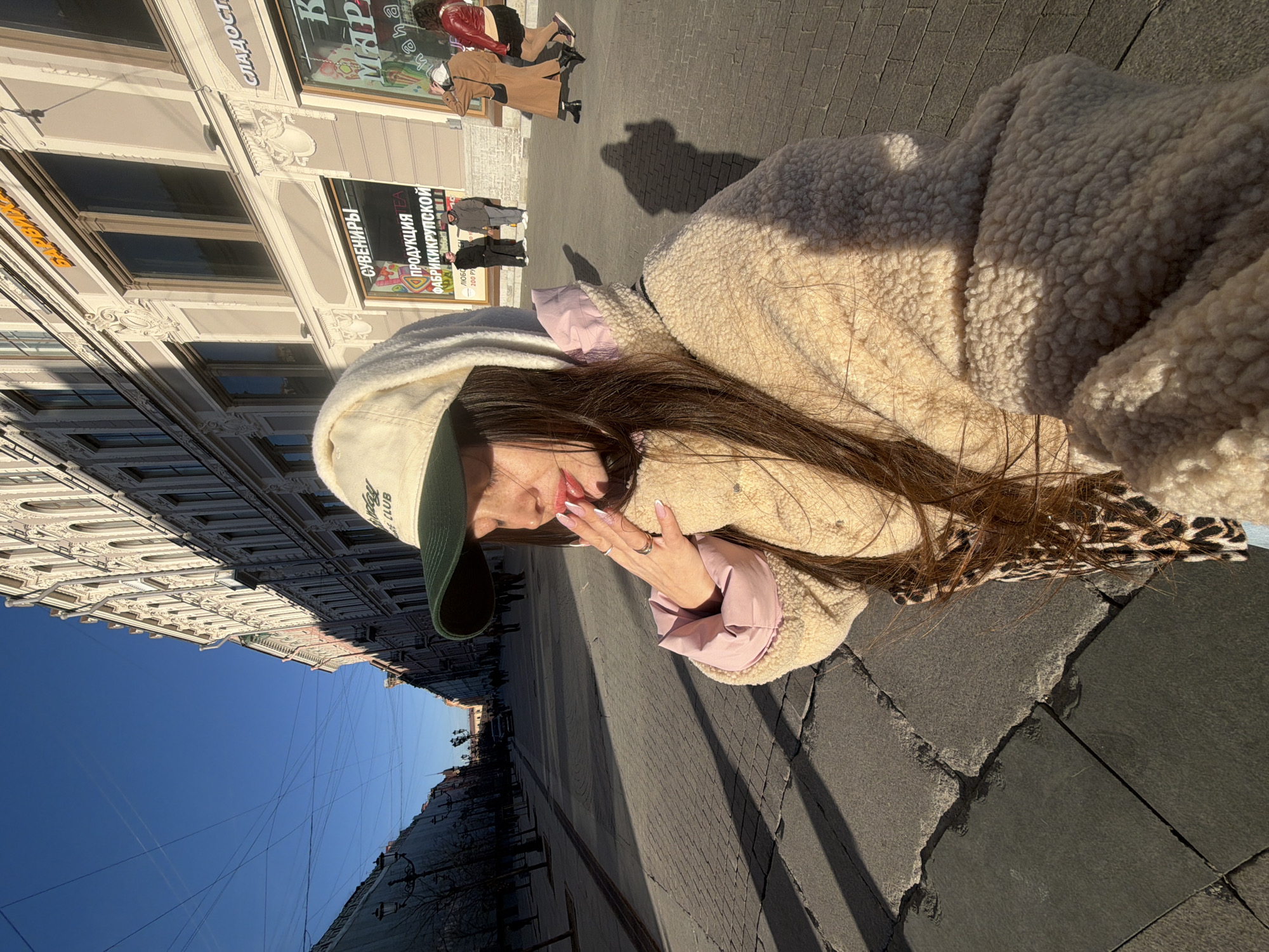 A woman stands on a sidewalk in Saint Petersburg, Russia. She wears a cream-colored, sheep-fleece jacket over a pink top, a baseball cap, and an animal print scarf. She is facing left with her hand raised near her mouth. In the background, people walk along the street next to a multi-story building. The sidewalk is made of rectangular paving stones. The sky is clear blue.