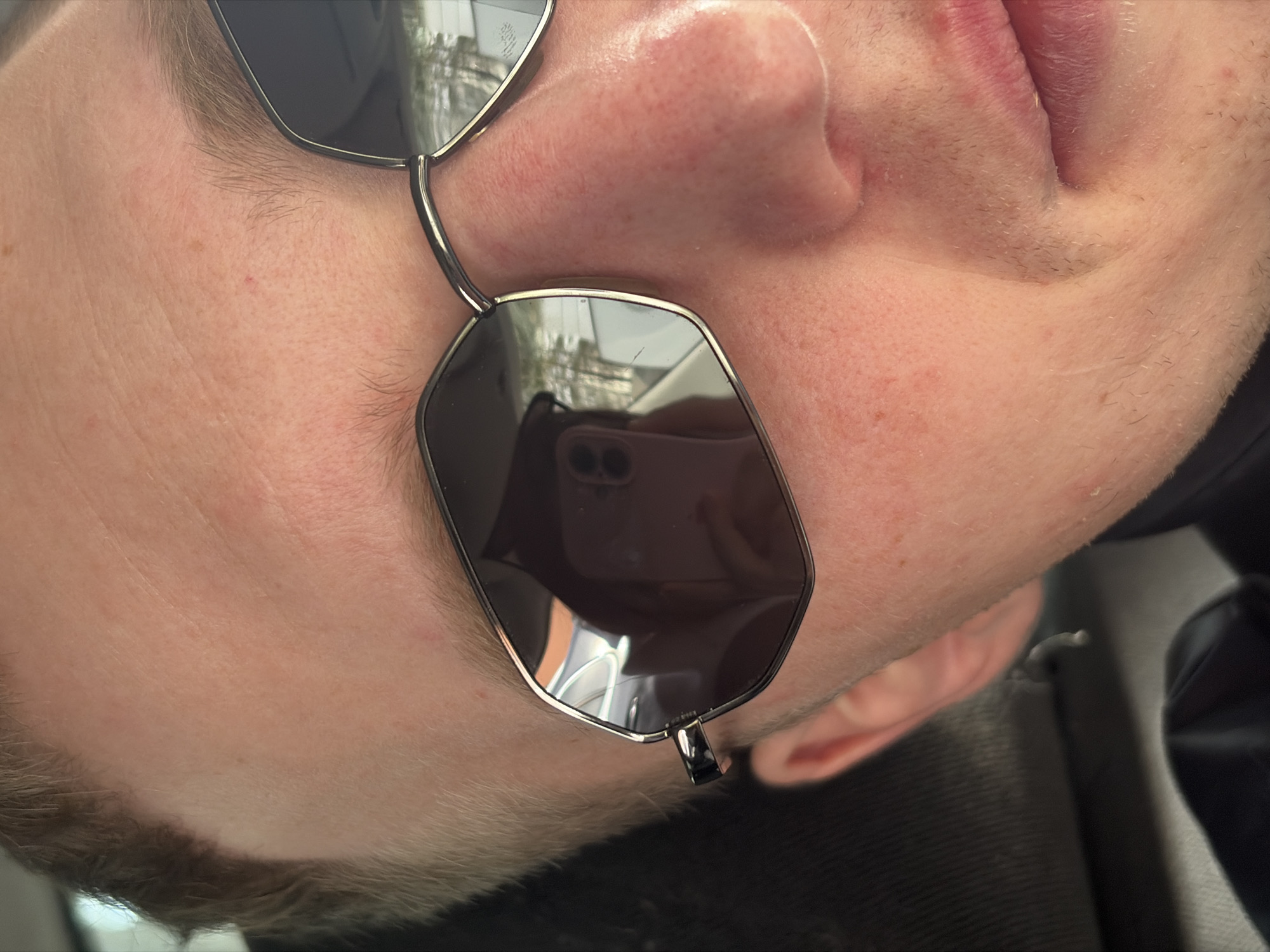 The image shows a close-up of a person's face wearing sunglasses. The reflection in the sunglasses reveals a pink phone. The person's skin is fair, and the image angle is slightly rotated. The location is not apparent from the image itself. Therefore, it is impossible to include details about the location in the city of Chaikovsky, Russia.