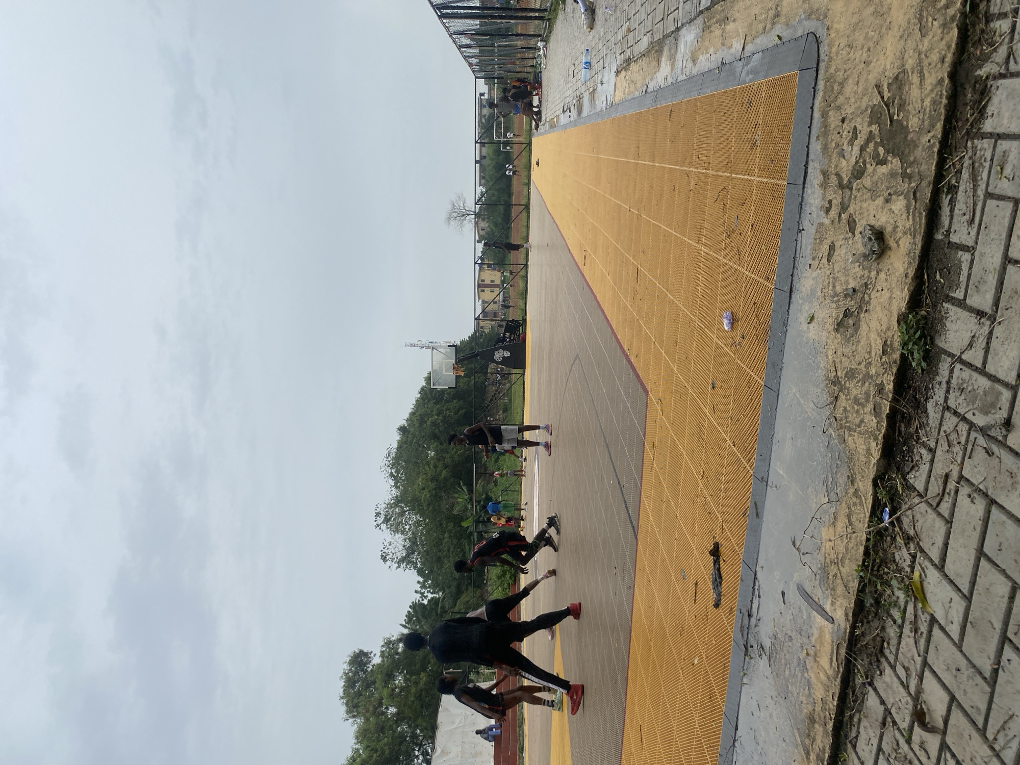 The image shows an outdoor sports court in Lagos, Nigeria. The court has a yellow playing surface with black perimeter lines and is surrounded by a gray concrete walkway. A chain-link fence borders one side of the court. Several young men are engaged in athletic activity on the court, some in running positions, others standing. Trees and buildings are visible in the background under a cloudy sky. Debris and foliage are scattered along the edges of the court and walkway.