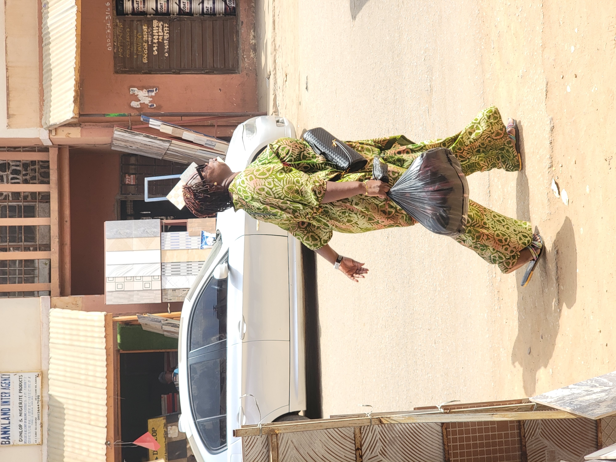 A woman in a green and patterned outfit and sandals walks along the sidewalk in Maiburiji, Nigeria. She is carrying a black purse and a black plastic bag. She is walking past a white car and a building with a shopfront. The shopfront has a sign that reads "BANKLAND INTER AGENCY" and advertises Dunlop & Nigerite Products. Tiles are stacked in front of the shop. The street is sunlit. A sign in the building above reads: "DAVIDO".