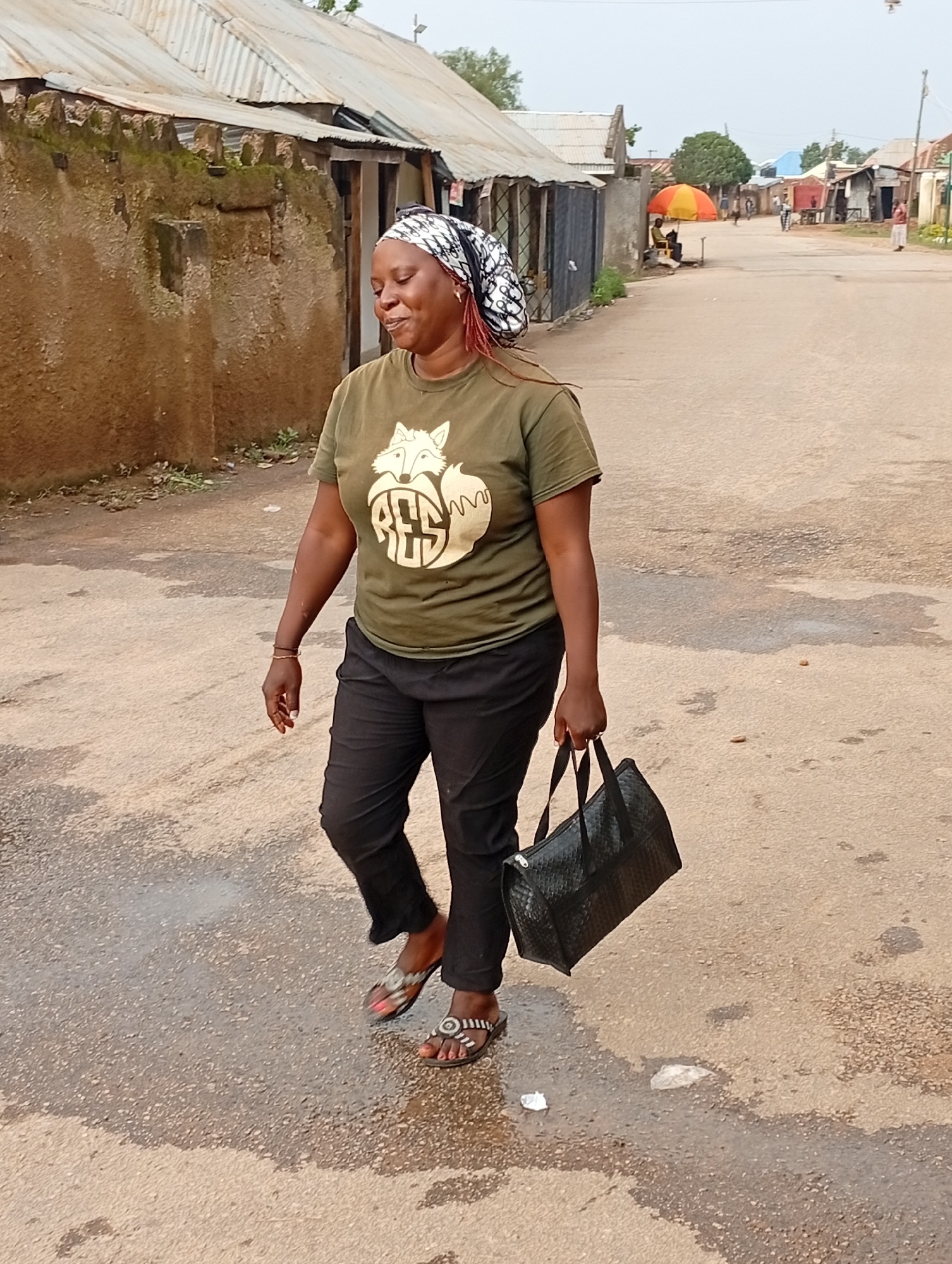 A woman is walking on a paved road in a residential area of Jos, Nigeria. She is wearing a green t-shirt with a fox graphic and the word "RED" in large letters. She is also wearing black pants and flip-flops. In her right hand, she carries a black woven handbag. The road is wet, suggesting recent rain, and there are puddles on the surface. To the left of the woman are buildings with corrugated metal roofs and mud walls. In the background, more buildings and a few scattered trees are visible. A person is seated under an orange and yellow umbrella on the right side of the road in the distance. The sky is overcast.
