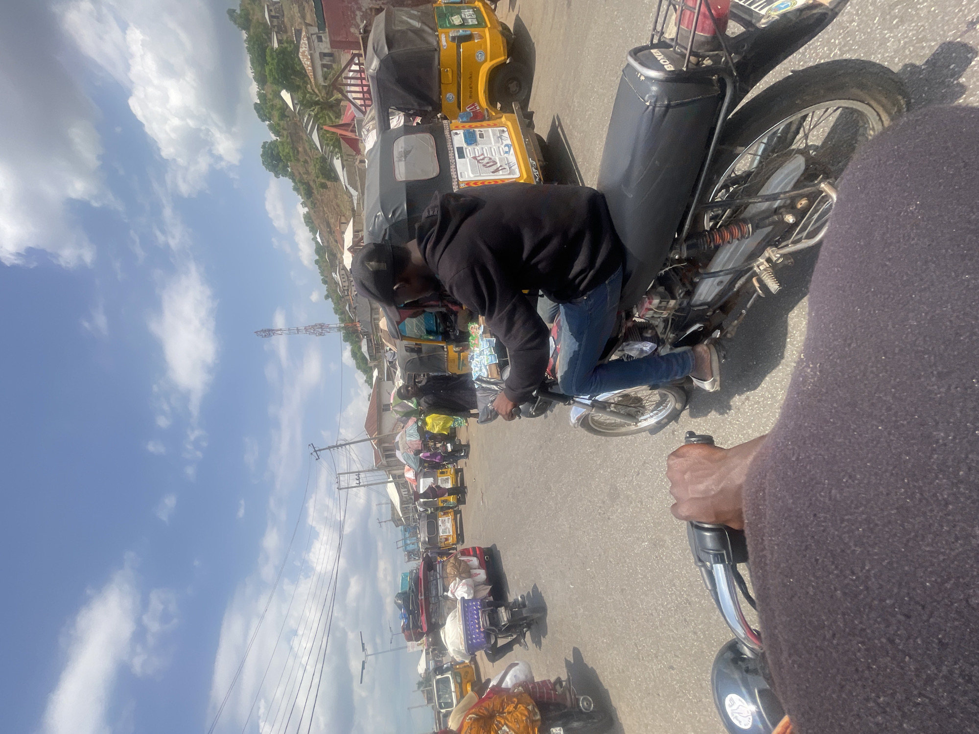 The image depicts a street scene in Lokoja, Nigeria, with multiple motorcycles and auto-rickshaws (locally known as "Keke Napep") visible. A person wearing a black hoodie and jeans is seated on a motorcycle. Another person, seen from the chest up and holding motorcycle handlebars, is closer to the camera. The street appears to be congested. In the background, buildings are visible on a hillside. The sky is partly cloudy.