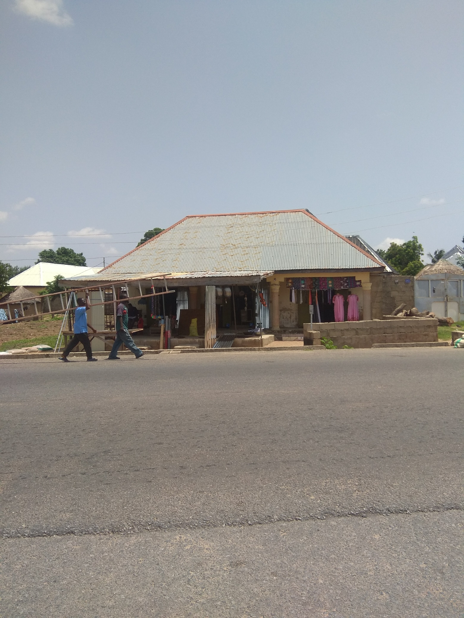 Two men are walking on an asphalt road, carrying a large wooden ladder between them. The man on the left is wearing a blue t-shirt and dark pants, while the man on the right wears a dark t-shirt and grey pants. They are walking from left to right in the frame. Behind them is a building with a corrugated metal roof, which appears to be a shop. The shop has its goods displayed outside, including some pink dresses hanging on the front. The building is made of concrete blocks and has a covered entrance. To the left of the shop, there's another building with a thatched roof, and further in the background, trees and a power line are visible. The sky is pale blue with a few wispy clouds. The scene is likely in Jalingo, Nigeria, given the architecture and general environment.