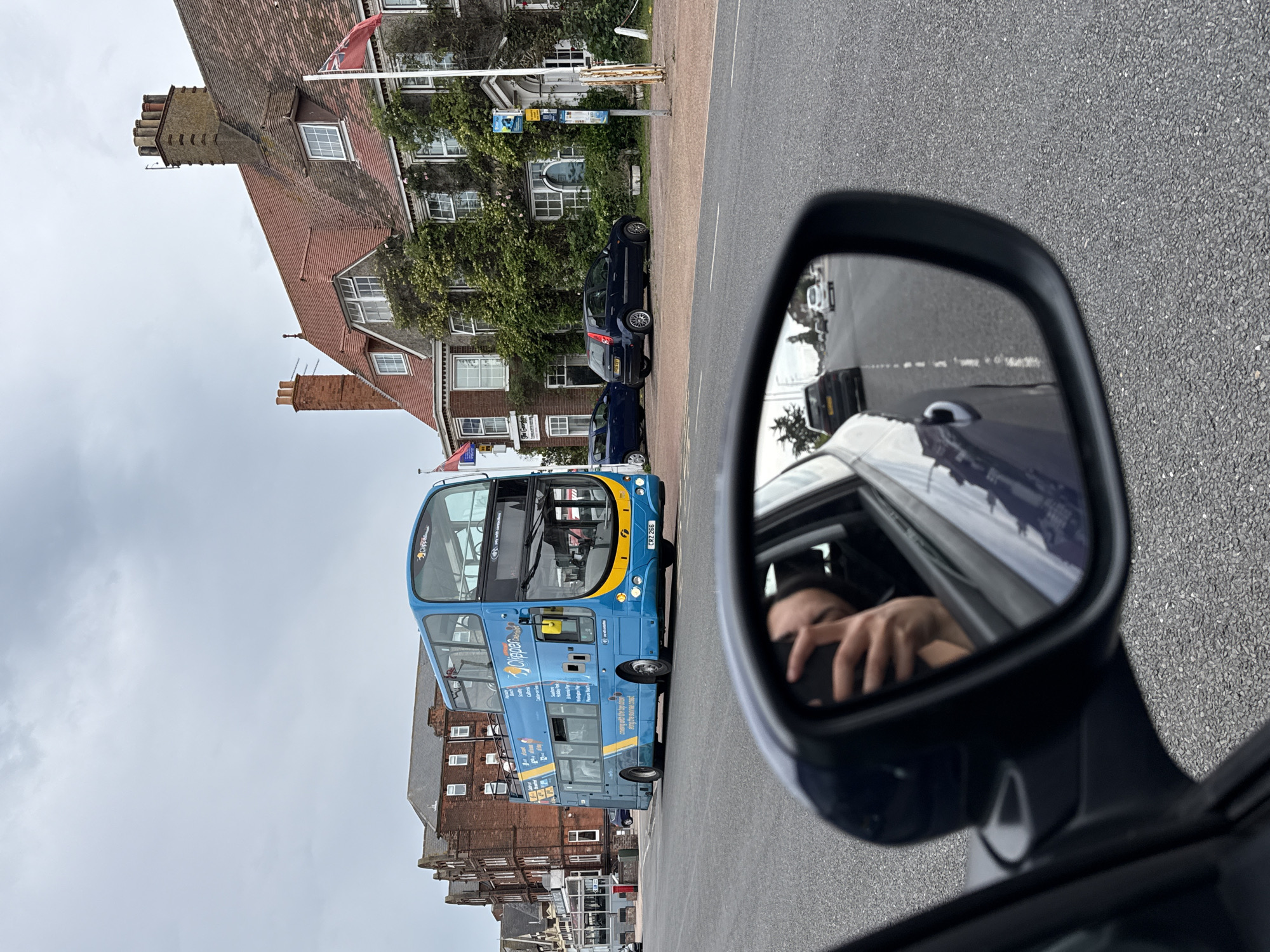 The image captures a street scene in Great Yarmouth, United Kingdom. A blue double-decker bus with "Clipper" branding is the dominant subject, traveling on a road. Adjacent to the road, on the right, is a red-tiled building, possibly a hotel or large residence, covered in ivy. Red flags are visible flying from the building. On the left side of the street, there are other brick buildings. In the foreground, a car's side mirror reflects the scene, including a person holding a smartphone, presumably taking the photograph. Several other cars are also present on the road. The sky is overcast.