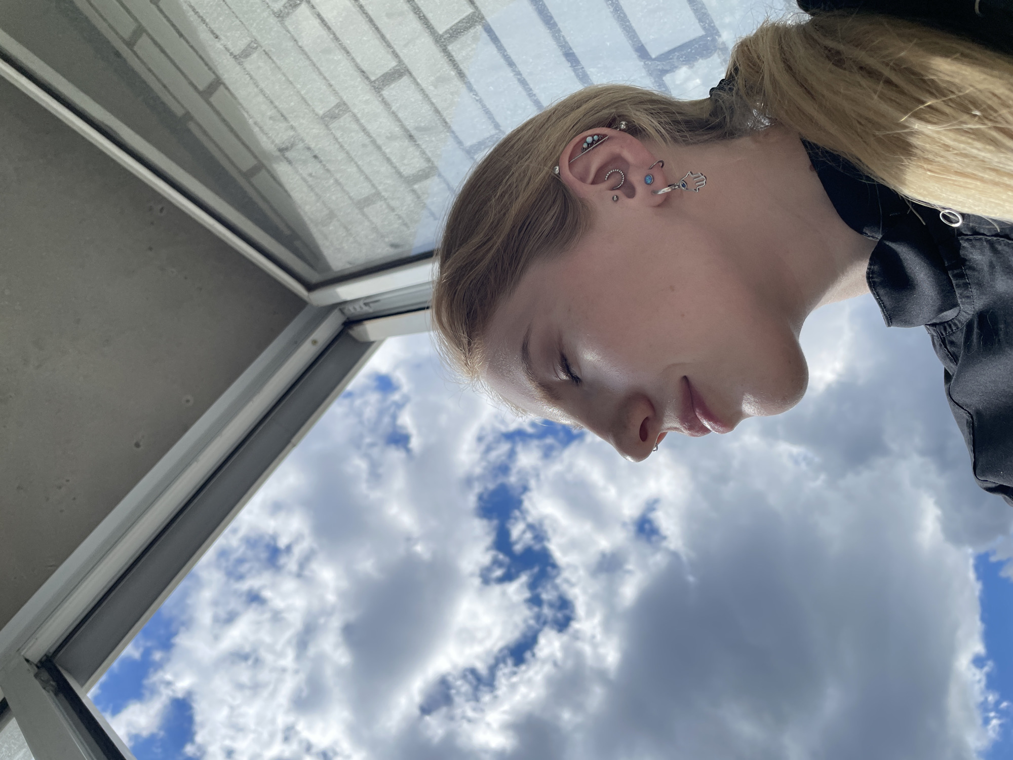 A blonde-haired person with numerous ear piercings is positioned in front of a window with a view of a partly cloudy sky. The person is wearing a black garment. The exact location within Izhevsk, Russia cannot be determined from this image.