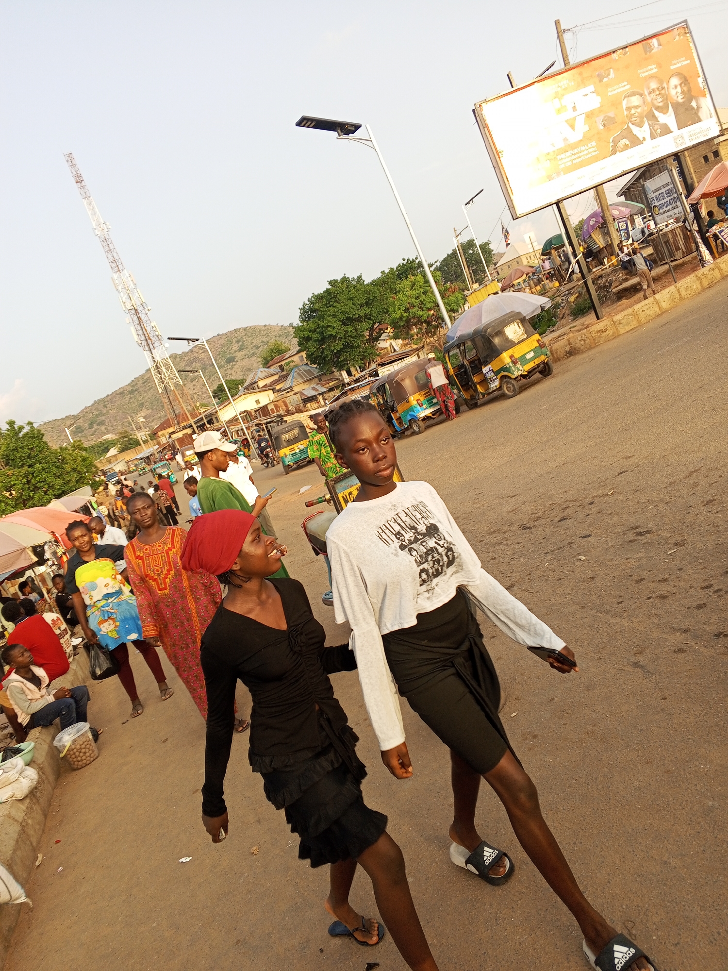 The image shows an outdoor scene in Gwong, Nigeria. Two young women walk on an asphalt street towards the right of the frame. The woman in front wears a long-sleeved cropped white shirt with a graphic design and a black skirt tied at the waist, along with Adidas sandals. The woman behind her wears a long-sleeved black dress, a red head wrap, and sandals. Other people are visible on the left side of the street, including vendors and other pedestrians. Three-wheeled vehicles (keke napep) are visible further up the street on the right. A large advertising billboard is positioned on the right side. A tall telecommunications tower stands in the distance on the left, and a small mountain range is visible in the background. Overhead street lights illuminate the area.