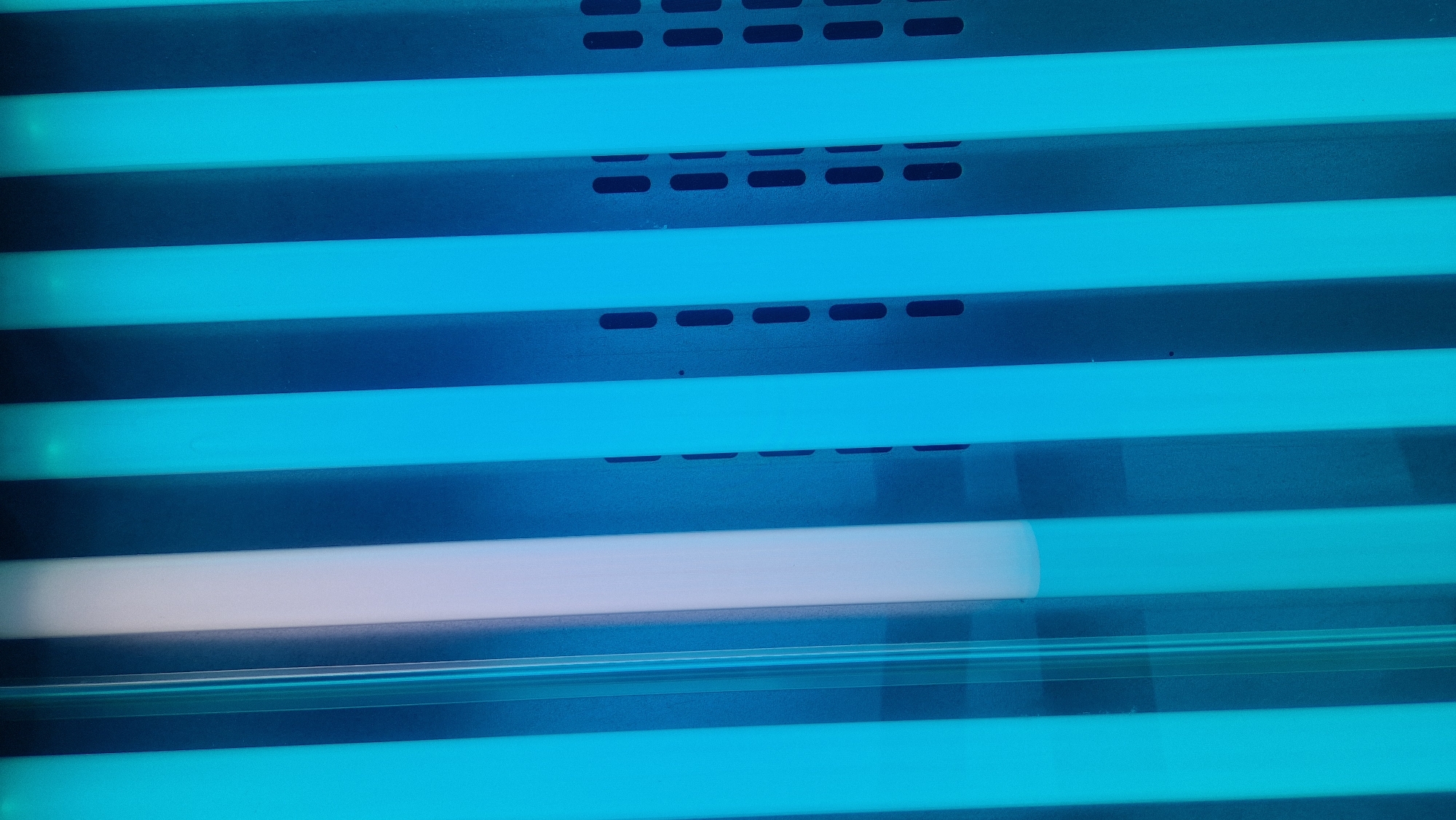 A vertical close-up of a tanning bed shows multiple blue fluorescent tubes and one white tube. The tubes are arranged in a parallel, slightly recessed manner, with visible ventilation slits on some of the blue tubes. The lighting is predominantly blue, with the white tube offering a contrast. The image appears to be taken inside a tanning salon. There are no people or discernable actions occurring in the scene. The setting suggests an indoor environment, likely a commercial establishment.