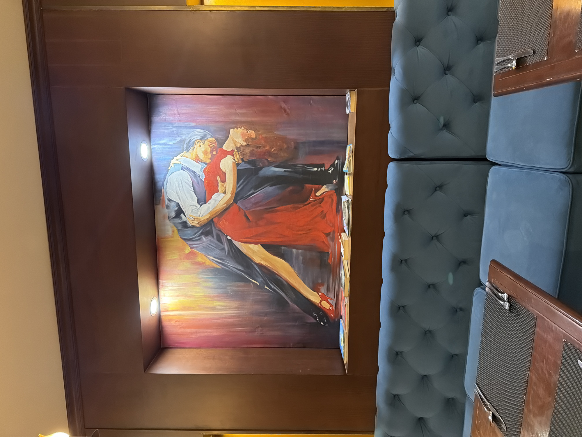 The image shows a painting of a couple dancing the tango, displayed in an alcove above a blue tufted sofa in what appears to be a restaurant. The painting features a man in a dark suit and a woman in a flowing red dress, their bodies contorted in a dramatic pose. The background of the painting is a vibrant, abstract blend of colors, primarily reds, yellows, and blues. The alcove has spotlights illuminating the artwork. Below the painting, on a wooden shelf, are several books. In the foreground, a wooden table with a dark placemat and cutlery is visible, suggesting a dining setting. The overall ambiance is one of elegance and artistry. The location is identified as Thuận Hóa Ward, Vietnam.