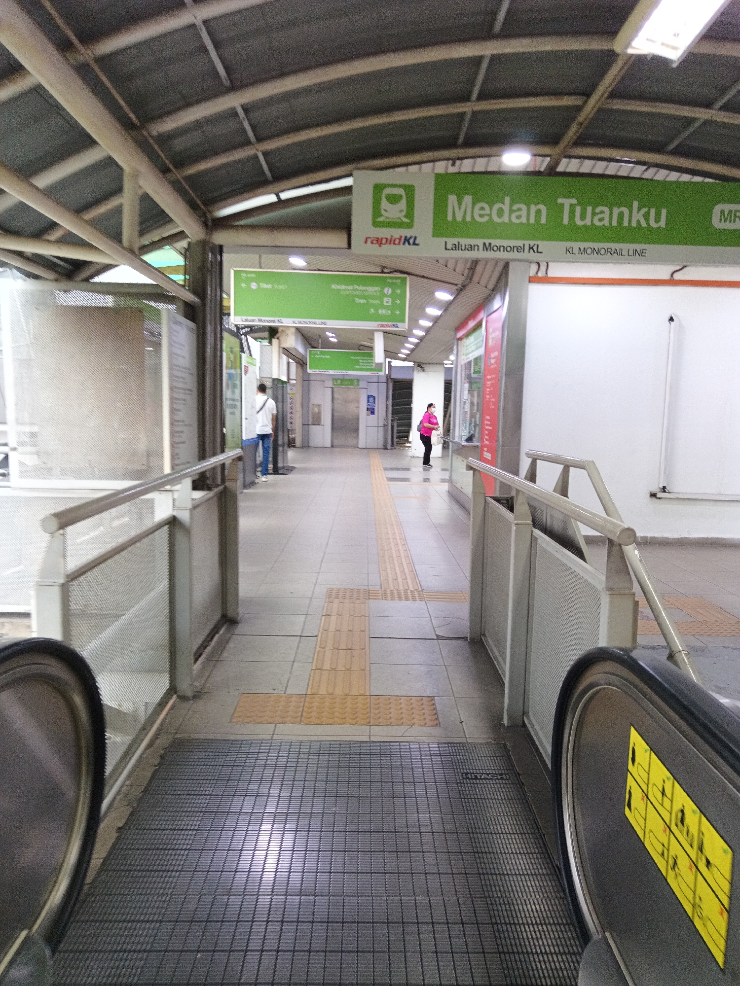 The image shows an interior view of the Medan Tuanku Monorail Station in Kuala Lumpur, Malaysia. The station features a covered walkway with a curved, corrugated metal roof supported by metal beams. A wide escalator, with the "HITACHI" brand visible on its surface, is present in the foreground on the left, and another escalator is partially visible on the right. The floor is tiled with a pattern of grey and yellow tactile paving. Signage indicating "Medan Tuanku" and "KL MONORAIL LINE" in green is visible on the right side of the walkway, with the RapidKL logo also present. Two individuals are present in the background. On the left, a person wearing a white shirt and blue pants is walking away from the camera. On the right, a person in a pink top and black pants is standing facing towards the right. The environment appears to be clean and functional, typical of a public transportation hub.