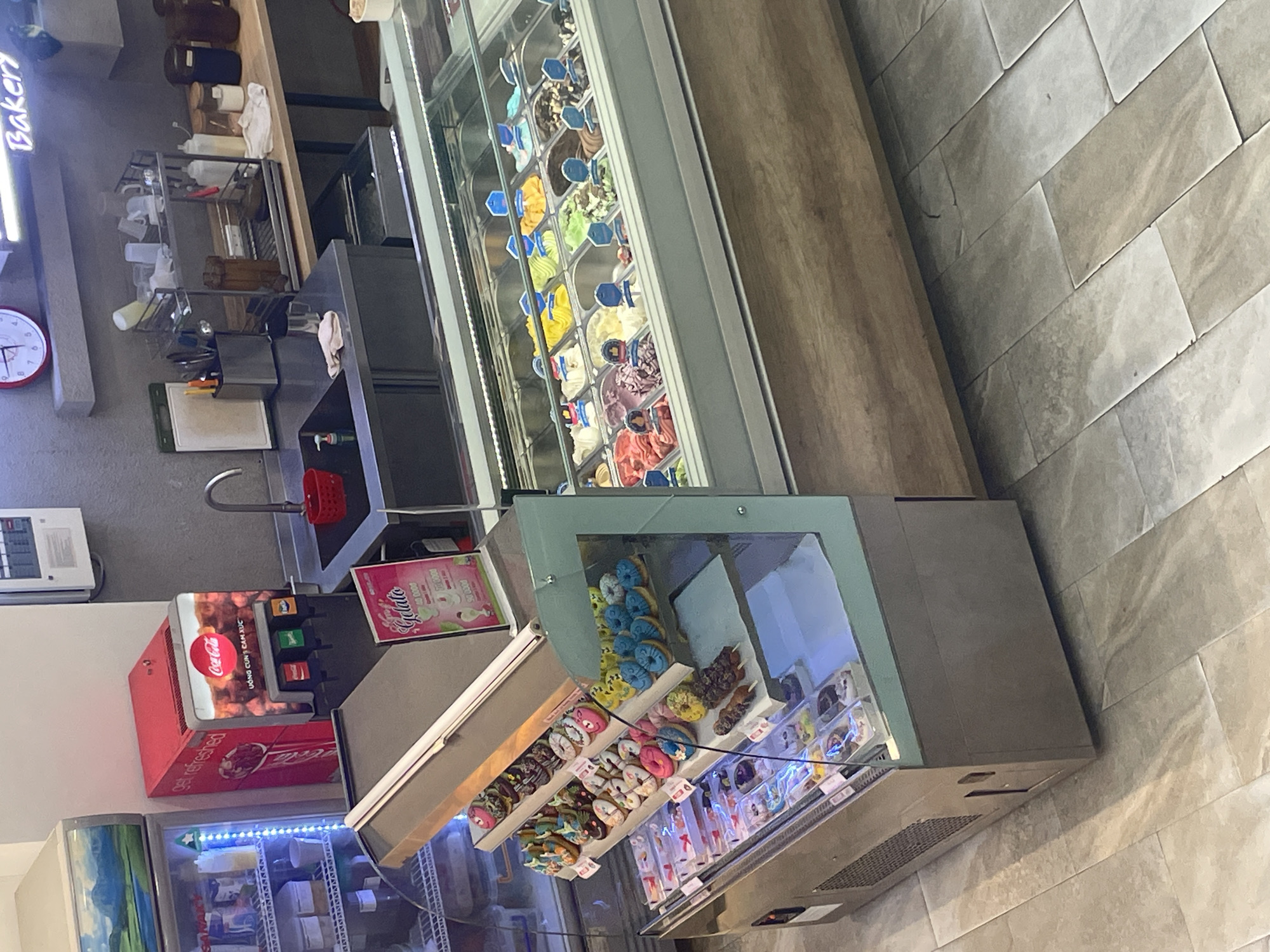 This photograph, taken in Nha Trang Ward, Vietnam, presents an indoor scene featuring food displays and service areas typical of a bakery or confectionery shop. The view is angled towards the right, showing various types of refrigerated display cases stocked with items such as gelato, pastries (particularly donuts), and bottled drinks.
The topmost display is a long, refrigerated case filled with various flavors of gelato, each labeled with a small sign. Below this, there is a refrigerated display case containing an assortment of colorful donuts and other pastries. A Coca-Cola dispenser is visible next to the donut display, and a refrigerated unit containing bottled drinks is situated beneath it.
In the background, a sink area with a faucet and a set of shelves stocked with various food preparation supplies and containers can be seen.