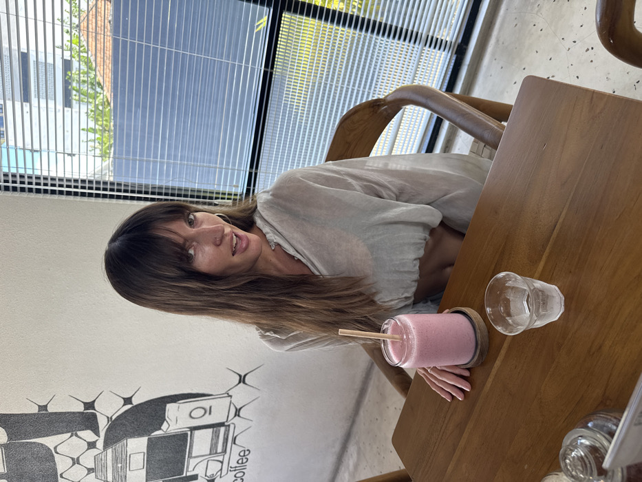 A woman with bangs and long brown hair sits at a wooden table near a wall with black and white art. She is wearing a light-colored blouse. On the table is a pink drink with a straw, a small empty glass, and what appears to be a menu. Behind her is a large window with blinds, letting in sunlight. She looks toward the camera.