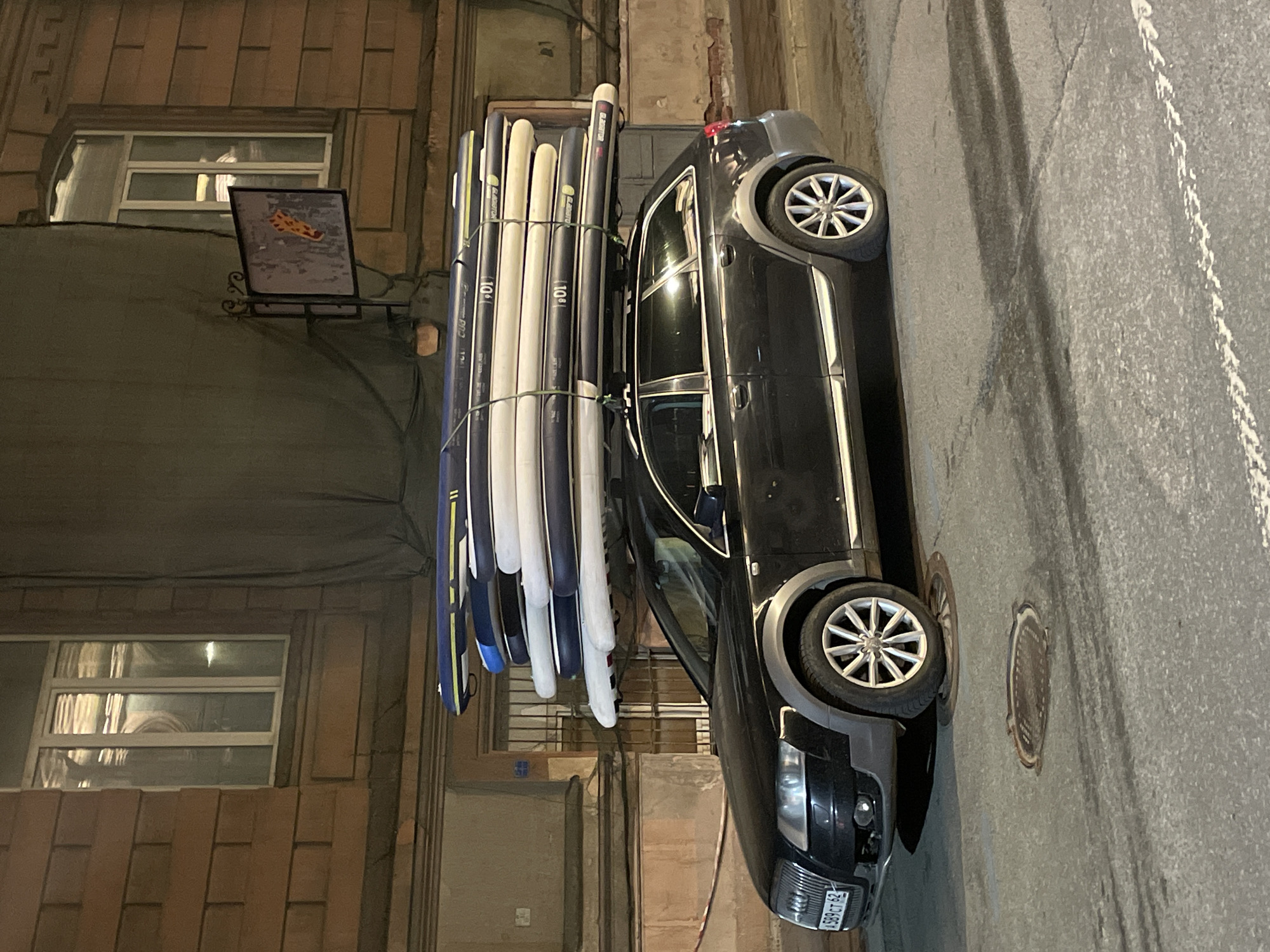 A black car is parked on a street in Saint Petersburg, Russia. It has a large number of paddleboards strapped to its roof. The license plate number on the car is "T 62". The street is paved and there is a manhole cover near the car. The buildings on the street are old and made of brick. There is a sign hanging above the street near the paddleboards that shows what looks to be a picture of pizza.