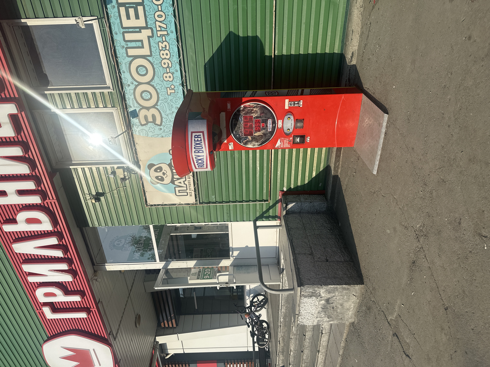 The image depicts the exterior of a store in Novoaltaysk, Russia. A red "Rocky Boxer" arcade game machine is situated on the pavement in front of a green corrugated metal wall. Above the machine, a sign for a pet store ("ЗООЦЕНТР") is visible, featuring a cartoon panda. To the left, there is an entrance to a store with the name "ГРИЛЬНИЦА" (Grillnitsa) displayed in large white letters on a red background. A bicycle is parked on the steps leading to the entrance, and the store's operating hours are posted on the door. The scene appears to be bathed in sunlight, casting shadows on the ground.