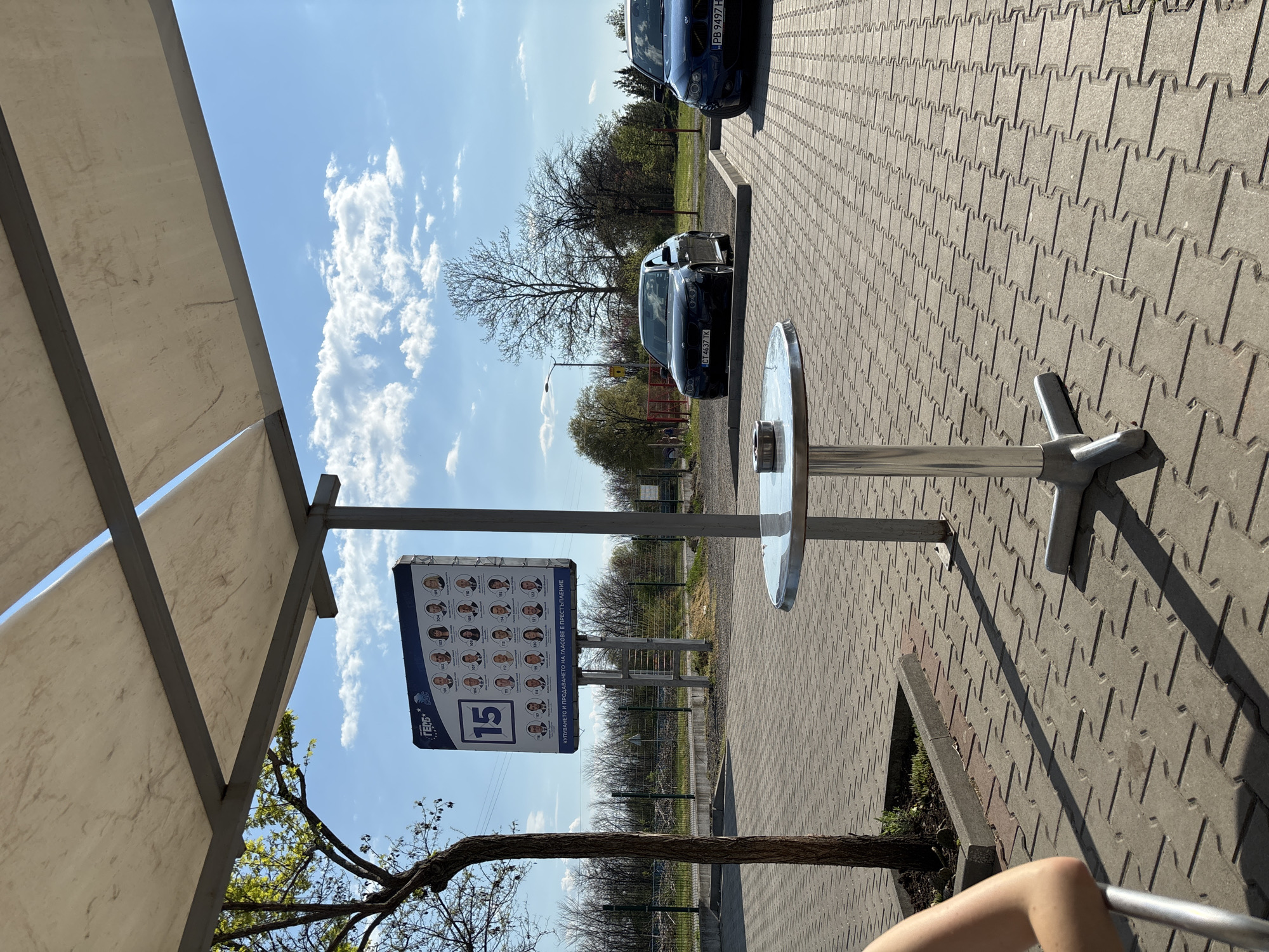 The image shows a parking area in Chirpan, Bulgaria on a sunny day. A metal table with a round top and a four-legged base is prominently placed in the foreground. A sign with portraits and the number 15 is mounted nearby, under the edge of a white canopy. In the background, several dark-colored cars are parked. The ground is paved with interlocking concrete bricks. The sky is blue with scattered clouds. A fence borders an area with trees. A small portion of an arm is visible in the bottom right corner.