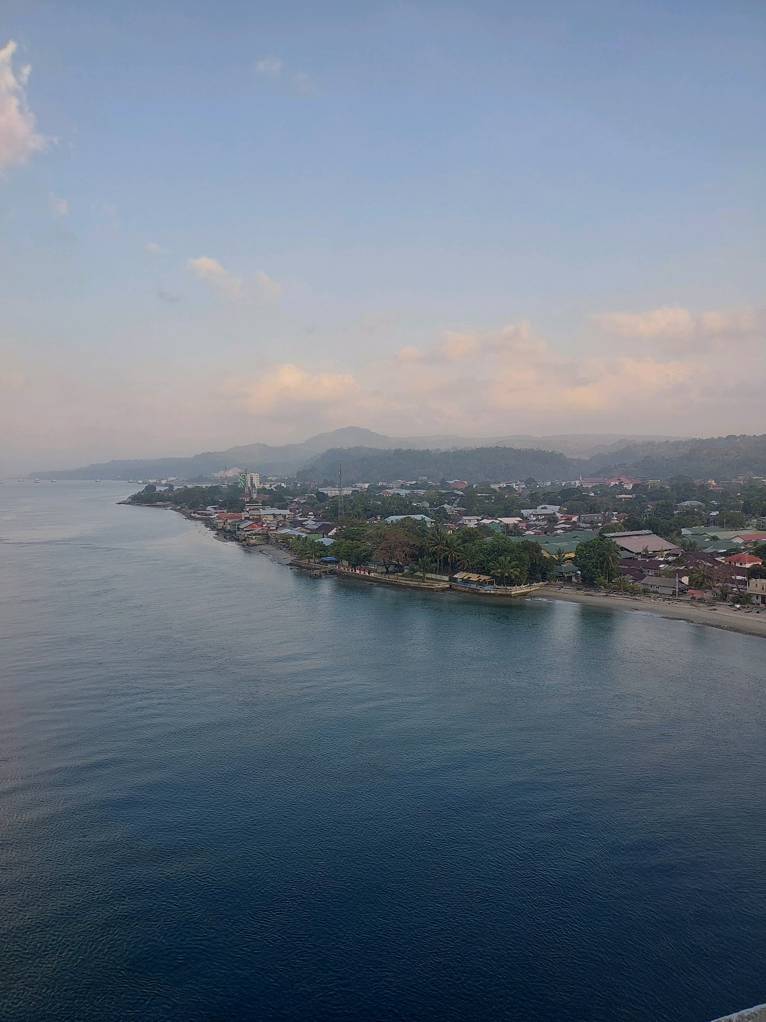 An aerial view captures a coastal settlement in Ambon, Indonesia. The scene unfolds along the water's edge, where a cluster of buildings with various colored roofs, some appearing to be homes and others shops, are situated. Dense green foliage, including palm trees, surrounds the structures, extending inland towards rolling hills that recede into the distance. The calm, dark blue water of the sea occupies the foreground and left side of the image, gently lapping against the shore. A thin, sandy beach is visible between the water and the buildings. The sky above is a pale blue, with sparse clouds scattered across it. The overall impression is one of a tropical coastal community bathed in soft, early morning or late afternoon light.