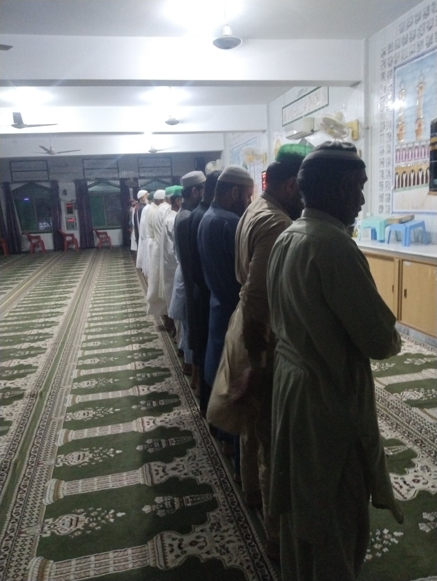 The image captures a group of men performing congregational prayer (Salah) inside a mosque or prayer hall in Dera Chahal, Pakistan. Approximately ten men are visible, standing in a straight line (saff) on green prayer carpets, facing an unseen qibla. Most of them are seen from behind or the side, wearing traditional Islamic attire including kurtas and skullcaps (topis). Their posture indicates they are in the standing phase of prayer.
The setting is an indoor space with a white ceiling featuring multiple fluorescent lights and several ceiling fans, one of which has four blades. The floor is covered with long, green prayer carpets patterned with light-colored motifs resembling mihrabs, columns, or floral designs, guiding the alignment of the worshippers.
The walls are light-colored. On the right, a wall features decorative tiles or murals, one depicting minarets and what appears to be a mosque structure (possibly the Kaaba in Mecca). Below these murals are light brown cabinets with several blue plastic stools placed on top. Further in the background, to the left, red chairs are visible against a darker, possibly windowed, wall section. Artificial lighting from the ceiling fixtures illuminates the scene, suggesting it is either evening, night, or a dimly lit interior during the day. Visible text on the wall appears to be Arabic calligraphy within framed art, though not clearly legible. The overall atmosphere is one of solemn religious observance.