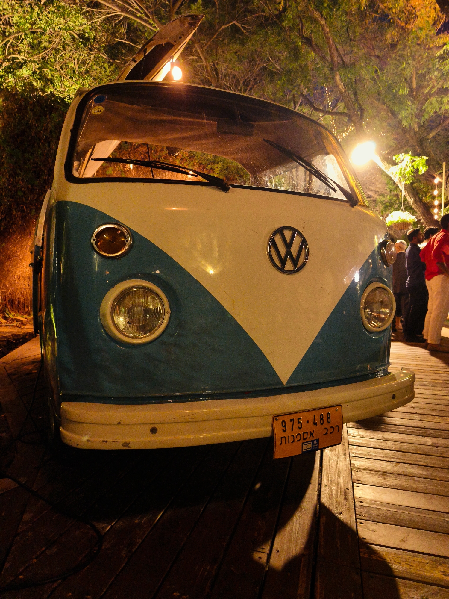 A vintage, two-tone blue and white Volkswagen Type 2 van is parked on a wooden deck at night, illuminated by artificial lighting. The van's hood is partially open, revealing a light source. The license plate, orange with black lettering, reads "975 488" and includes Hebrew text. Several individuals are visible in the background, some standing and some seated, on a raised platform or stage. The environment suggests an outdoor event or gathering, with trees and string lights visible in the background. The overall scene is lit with warm, yellow and orange tones from the artificial lights. The location is specified as Hulda, Israel.