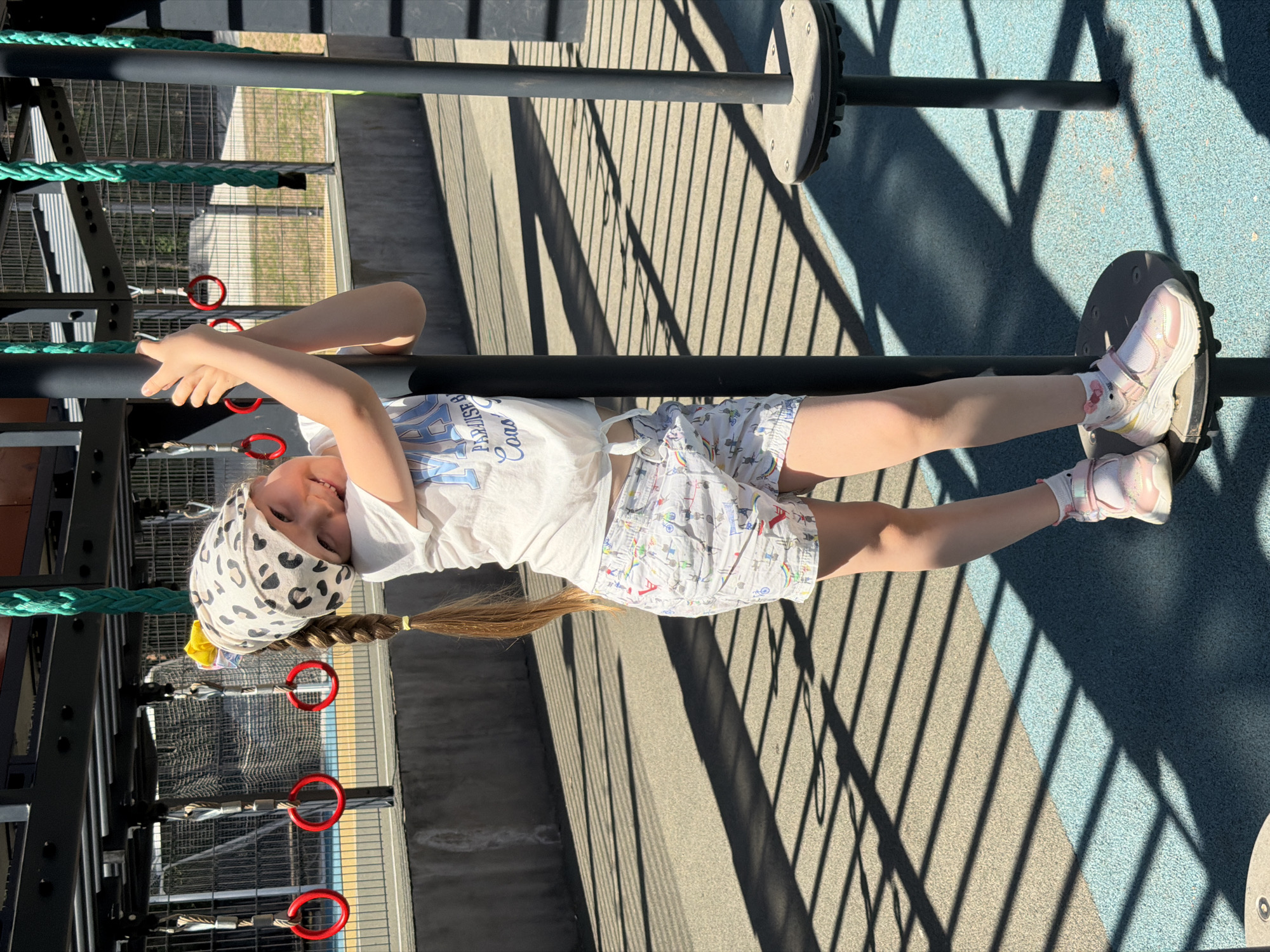 A young child is captured in a vertical shot taken outdoors on a sunny day. The location appears to be a playground or recreational area, based on the presence of play equipment such as ropes, rings, and platforms. The girl is suspended horizontally between two bars and resting her feet on a platform. She is wearing a white t-shirt, patterned shorts, and white sneakers with pink accents. Her hair is styled in a braid with a yellow hair tie, and she is wearing a printed bandana. The ground is covered with a blue-gray surface, and there are shadows of the playground equipment cast across the scene. The location in the city of Izhevsk, Russia is not supported by the image data.