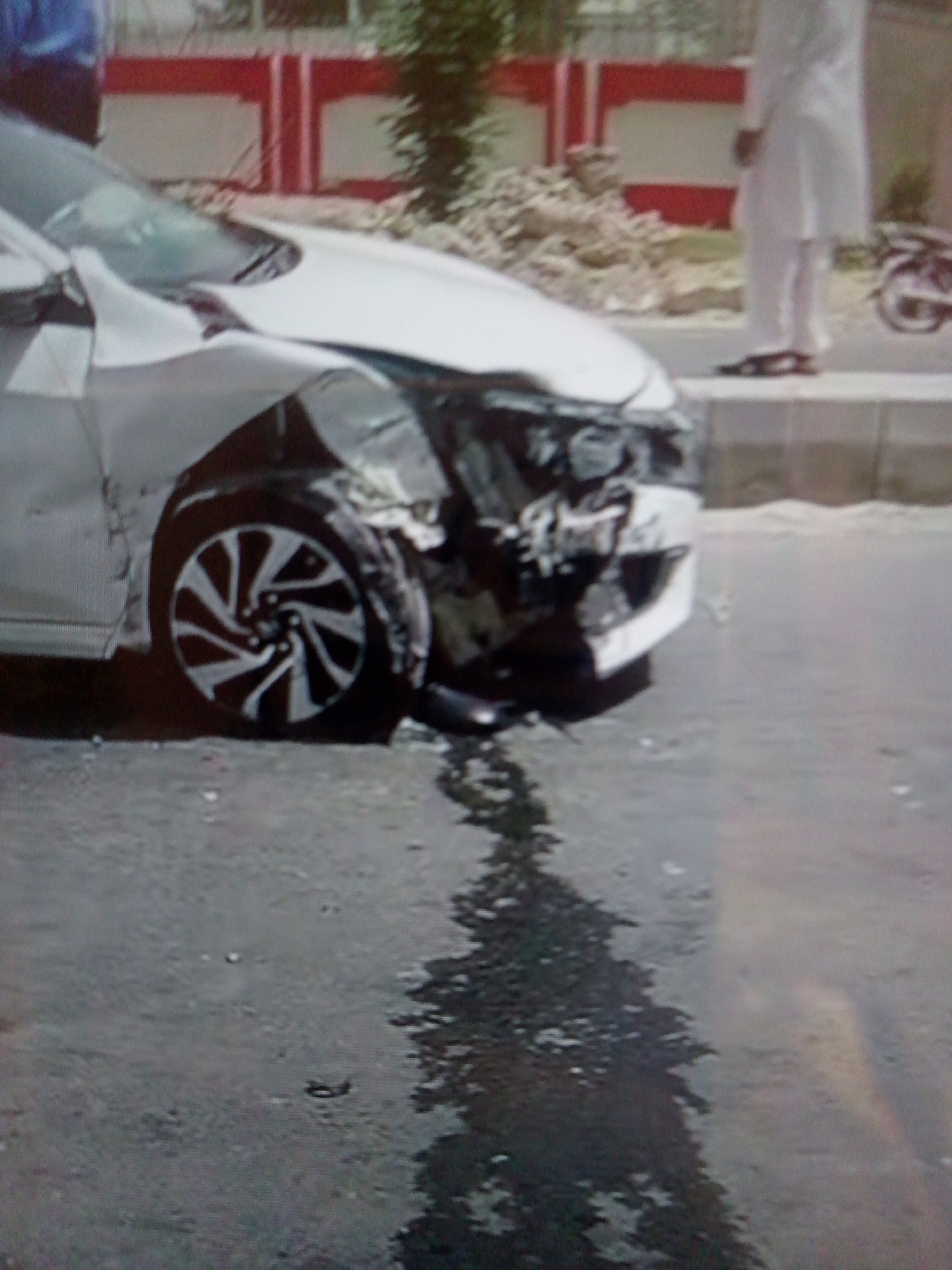 The image shows a damaged white car after an accident. The front of the car is severely damaged, particularly around the front left wheel. There is a wet surface, possibly from rain, with reflections. A man in white clothing stands on a sidewalk next to the car, and a parked bicycle is visible in the background. The location is a street, and in the background, there is a low brick wall with red and white accents. The city is unknown.
