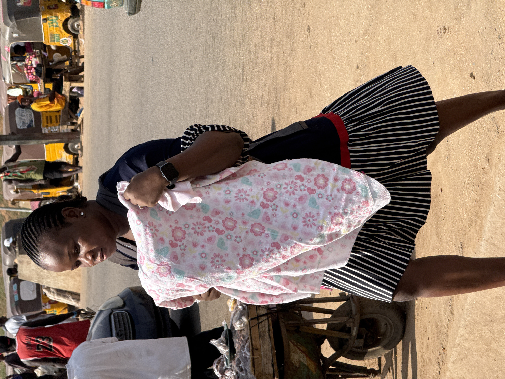 A woman walks along a paved street in what appears to be a busy market or urban area in Abuja, Nigeria, during bright daytime. She is the central figure, with dark braided hair and an attentive expression, looking downwards. She wears a dark blue top and a black and white striped skirt with a red waistband accent, and a smartwatch on her left wrist. She carries a large, folded bundle or blanket, predominantly pink and white, adorned with a repeating pattern of small flowers and hearts.
The background is bustling with activity. Several yellow motorized tricycles, commonly known as 'keke napep', are visible, some parked and others suggesting movement. Other people are present, some standing near what look like market stalls or small carts laden with goods. Part of a dark-colored car is also visible. One person in the background wears a red shirt with the number '23' printed on it. The bright sunlight casts distinct shadows, indicating clear weather. The overall scene depicts a typical busy street environment.