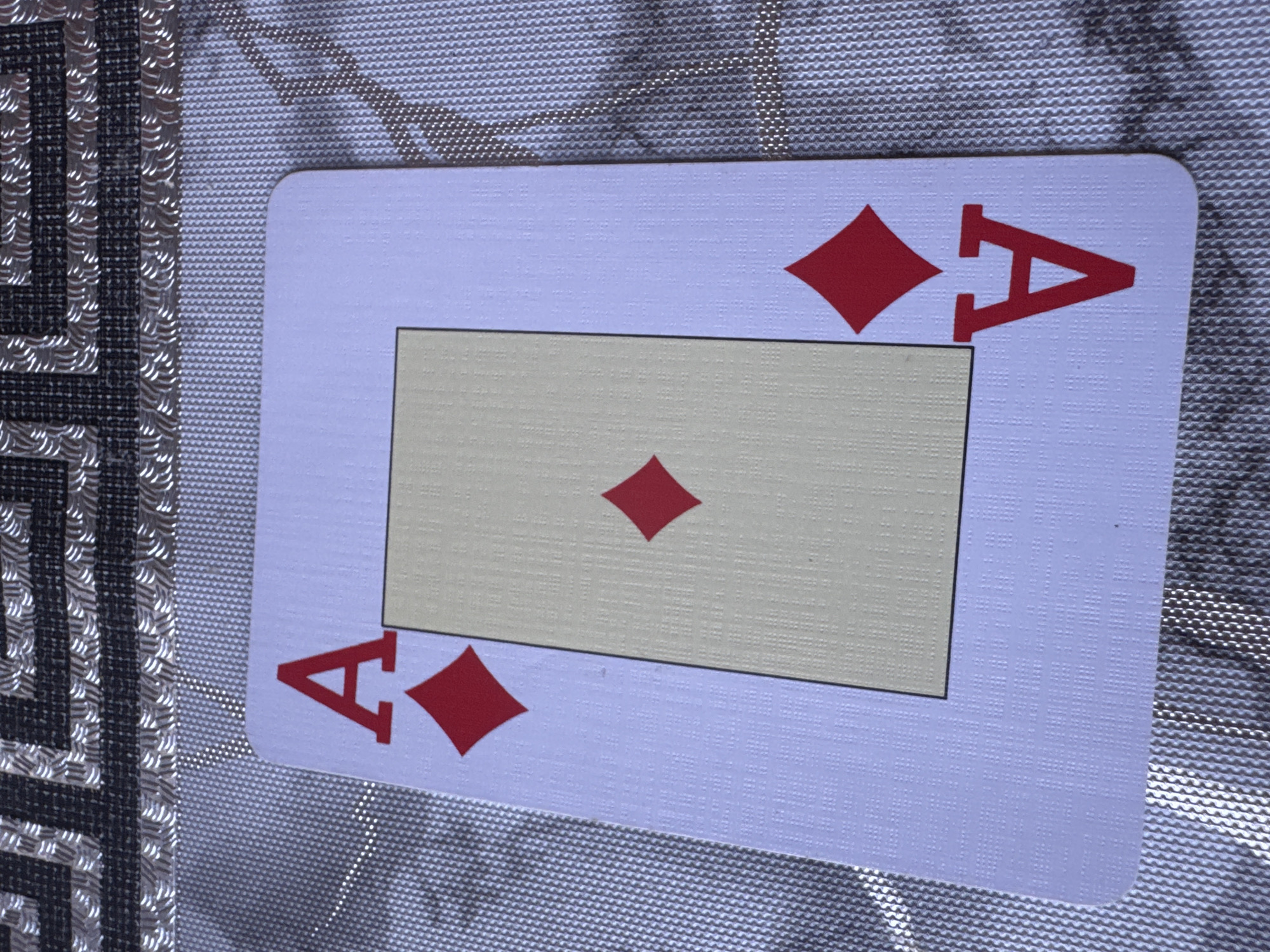 A close-up, overhead shot of an Ace of Diamonds playing card is positioned diagonally on a textured, reflective surface. The card is white with a light yellow rectangle in the center and red diamonds and the letter 'A' indicating its suit and rank. The background surface has a patterned texture with prominent silver thread accents, creating a shimmering effect. The image does not depict any city, people, or actions.