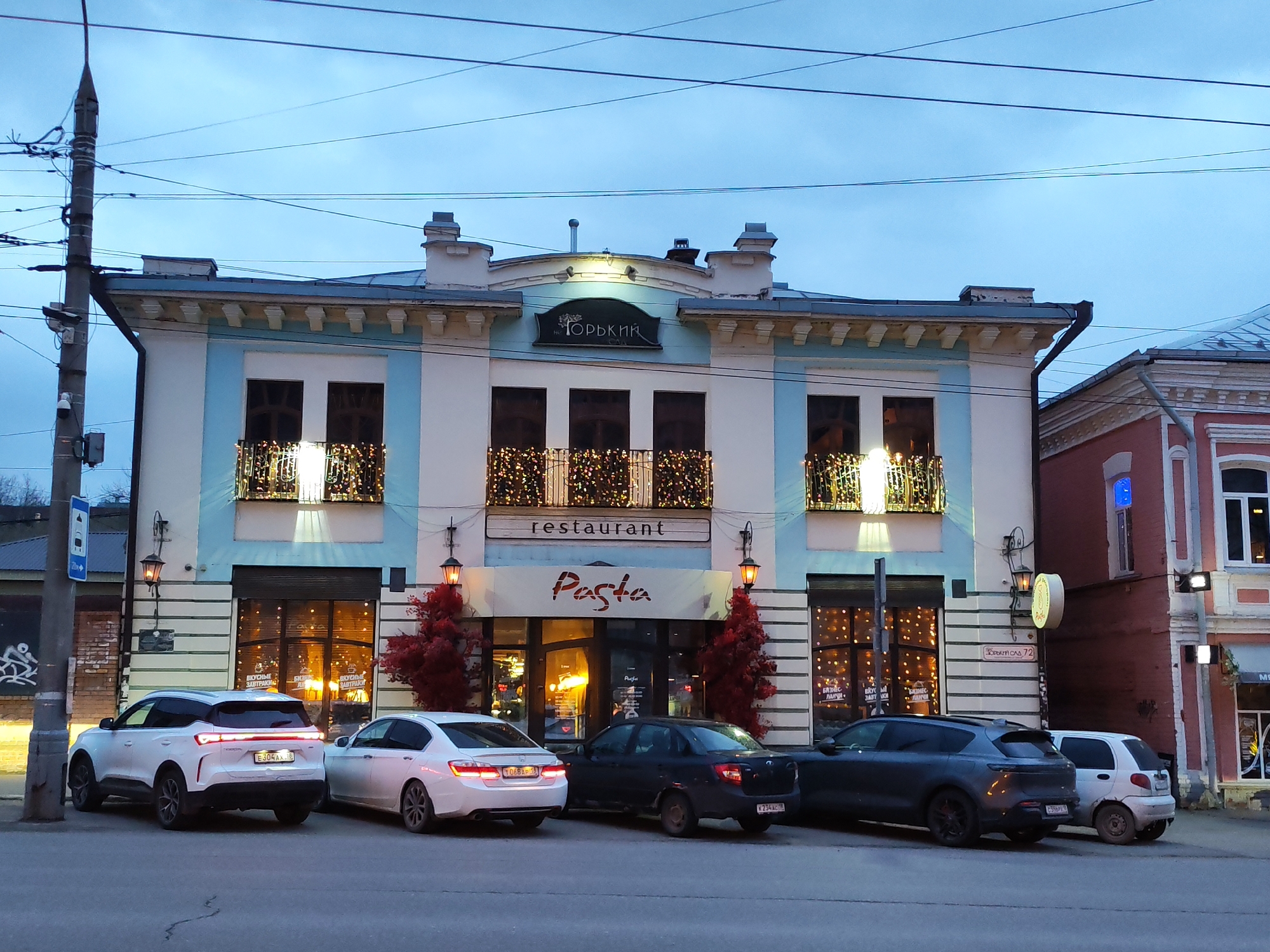 The image captures an evening street scene in Izhevsk, Russia, under an overcast sky, with artificial lights illuminated. The central subject is a two-story building with a light blue and off-white facade, featuring classical architectural elements and decorative cornices. The building houses a restaurant, prominent with festive string lights adorning its second-story balconies and ground-floor windows.
A sign at the top of the building reads "Горький" (Gorky), while a larger sign below the balconies simply states "restaurant" in English. An awning over the main entrance displays "Pasta" in a stylized script, with "Паста" also visible on the entrance door. Inside the windows, decals advertise "ВКУСНЫЕ ЗАВТРАКИ" (Delicious Breakfasts). Two artificial red trees flank the restaurant's entrance, adding a splash of color.
Several cars are parked along the street in front of the building. From left to right, a white SUV (license plate E304AA 18), a white sedan (T068AP 18), a dark sedan (X234XC 18), and a dark SUV (C599PY 18) are visible. All legible license plates bear the region code "18," consistent with the Udmurtia region where Izhevsk is located. To the far right, a small white car is partially visible.
On the left, a concrete utility pole laden with numerous overhead wires and a small security camera stands beside the road. To the right of the main restaurant building, a reddish-brown building with simpler architecture is partially visible, featuring a street sign that reads "Удмуртская 72" (Udmurtskaya 72), indicating its address. The scene appears to be a typical urban street during the early evening hours.