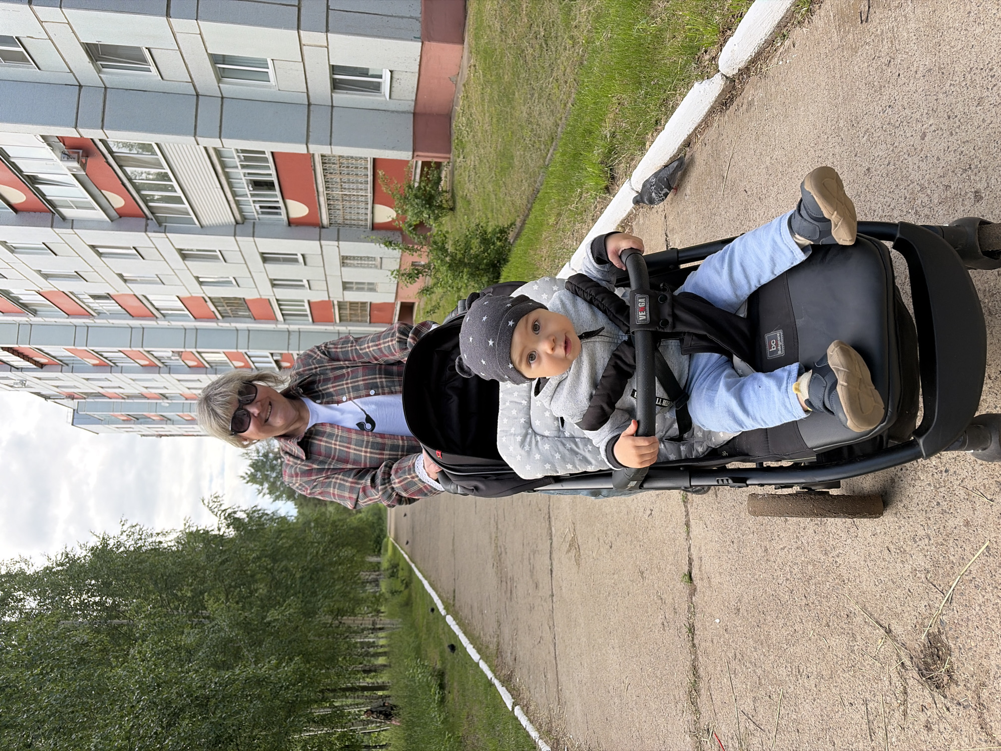The image shows a woman pushing a baby in a stroller on a paved path in Chaikovsky, Russia. The woman, with blonde hair and sunglasses, wears a plaid shirt over a white t-shirt and smiles warmly at the camera. The baby, likely a toddler, is seated in a black stroller, wearing a grey hooded outfit with white stars on the hat, light blue pants, and brown shoes. The baby looks towards the viewer with a curious expression, holding onto the stroller's front bar.
The scene is outdoors, with a concrete or asphalt path featuring a white painted line. Lush green grass and mature trees are visible alongside the path and in the background. A large, multi-story apartment building with a distinctive grey, red, and white facade, characteristic of post-Soviet residential architecture, rises behind the subjects. A pigeon is visible on the path near the stroller.
The activity is a casual outdoor stroll. The time of day appears to be daytime, possibly with an overcast sky, providing soft, diffused light. Visible text includes "VEGA" on the stroller's handle and "bc babycare" on the stroller's seat. The overall mood is pleasant and relaxed.