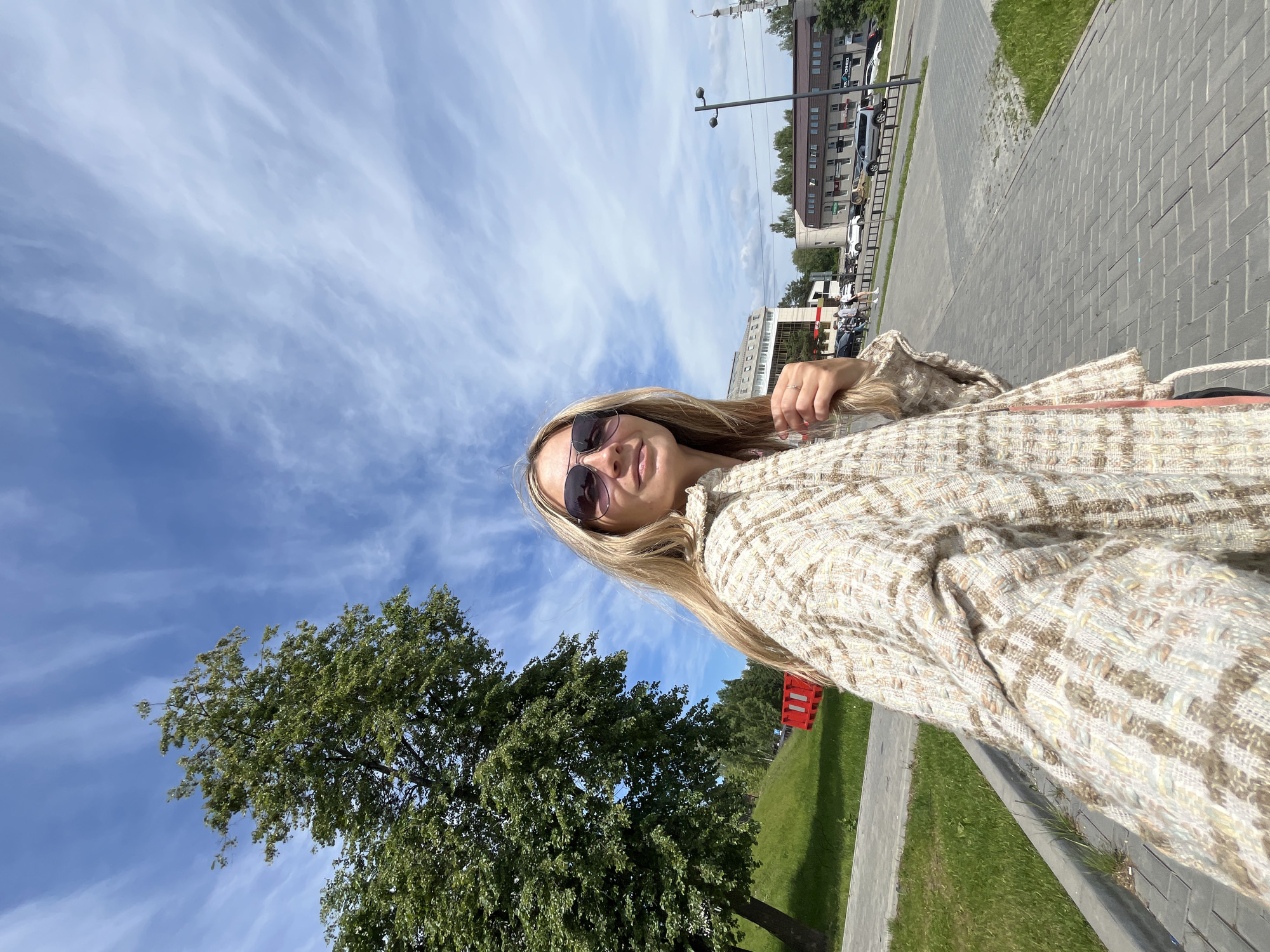 The image is a low-angle selfie featuring a young woman with long blonde hair, wearing sunglasses and a textured, light-colored jacket, possibly tweed or woven. She has a subtle smile or smirk on her face. The camera is angled upwards, capturing a significant portion of the bright blue sky, which is adorned with long, wispy white clouds, suggestive of cirrus clouds.
In the lower-left section of the frame, a large, lush green tree with dense foliage is prominent. The background on the upper-right reveals an urban setting, including several multi-story buildings, a street with parked cars, a paved pedestrian area, and a grassy verge. A streetlight pole and a partial view of a red barrier or container are also visible.
The scene depicts a sunny daytime setting in an urban environment, indicative of a street or park area in Chaikovsky, Russia. The overall impression is one of a casual outdoor moment. There is no discernible text visible in the image.