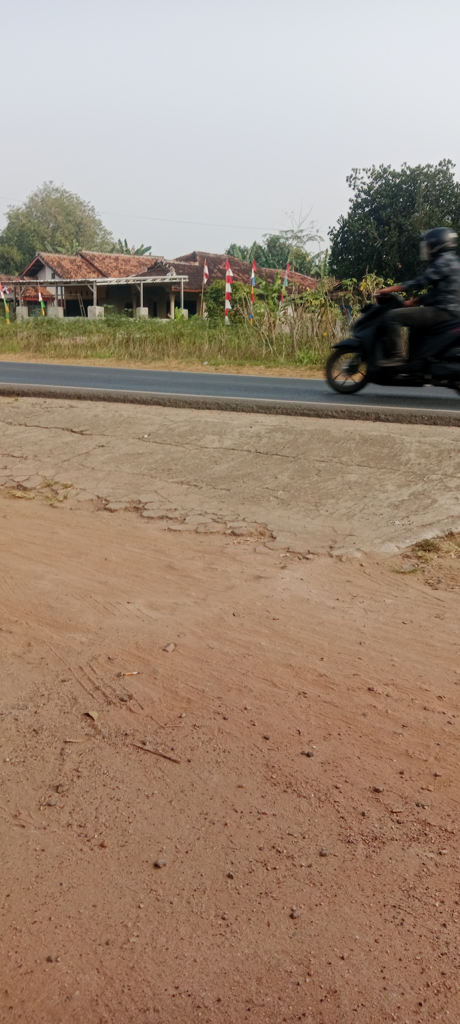 In Gondang Rejo, Indonesia, a blurry motion shot captures a person on a black motorcycle driving on a paved road. The motorcycle and rider are in motion, appearing as a dark silhouette. The road is bordered by dry, cracked earth on the foreground and a patch of overgrown grass on the right. In the background, a traditional Indonesian house with a reddish-brown tiled roof stands behind a line of trees. Several small Indonesian flags, predominantly red and white, are displayed in front of the house, suggesting a local celebration or event. The sky is overcast with a pale, hazy atmosphere.