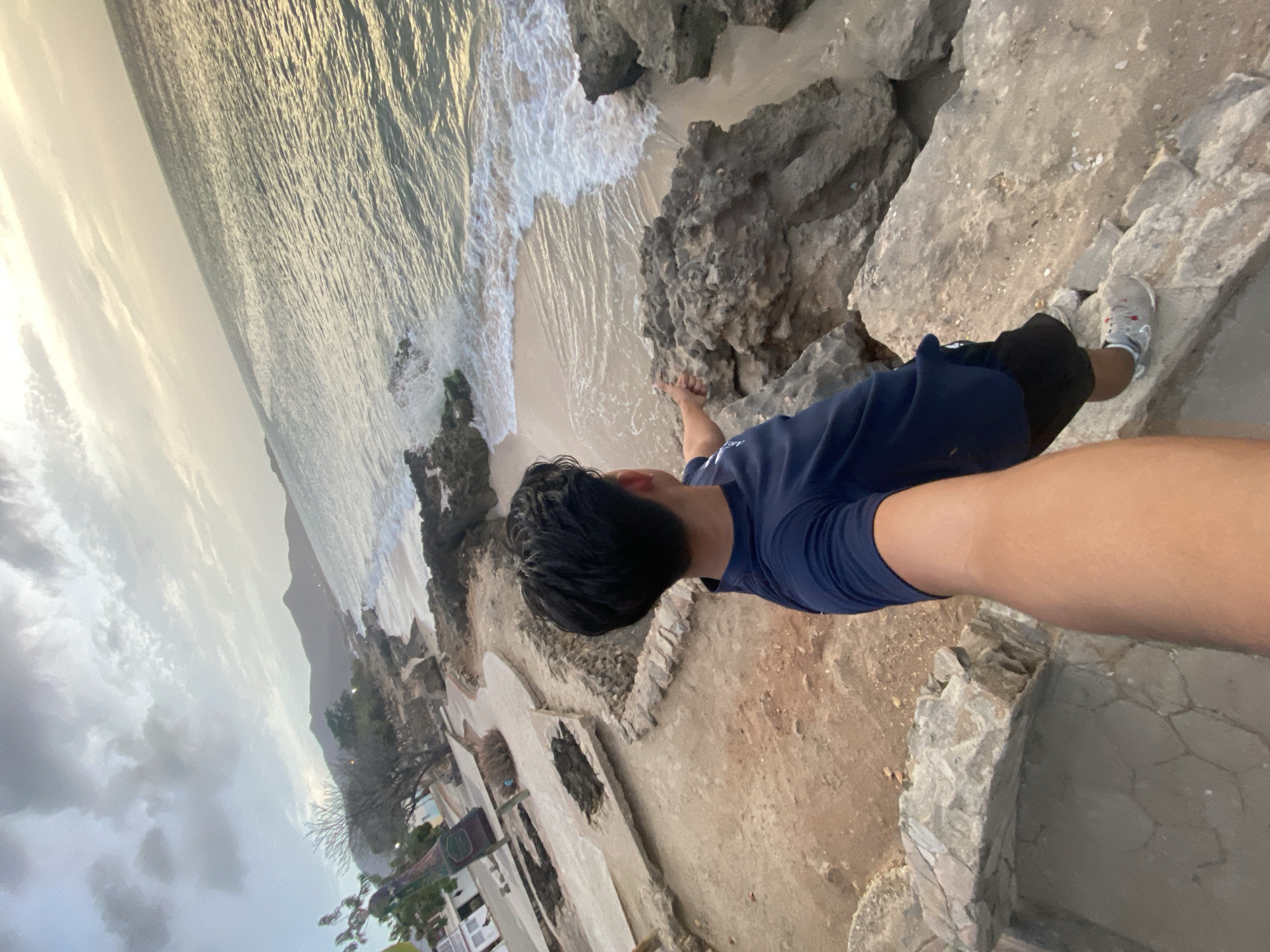 The image shows a person standing on a rocky precipice overlooking a beach. The location appears to be coastal, with a beach, ocean waves, and a cloudy sky. The person is wearing a blue shirt and black shorts, and they are standing near some concrete structures. The location is Juan Griego, Venezuela.