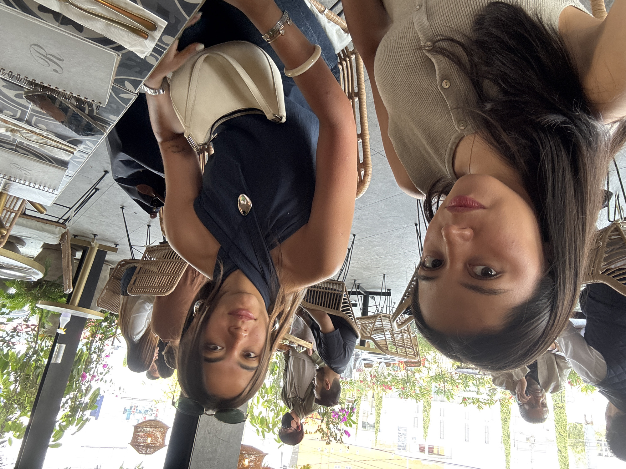 The image captures a vibrant scene inside a modern restaurant or cafe, taken from the perspective of two women facing the camera. In the foreground, two women are prominently featured. The woman on the left wears a dark blue top with a criss-cross neckline, possibly denim jeans, and accessorizes with a light cream-colored shoulder bag, a silver bracelet, a cream-colored bangle, and sunglasses perched on her head. The woman on the right is dressed in a light beige, ribbed cardigan-style top. Both women have dark hair and are looking directly at the camera with neutral to pleasant expressions.
They are seated at a table with a distinct dark and light patterned surface. On the table, two light-colored, spiral-bound menus are visible, each adorned with a stylized initial "R" on the cover. Cutlery, including forks and knives, is neatly wrapped in white napkins.
The restaurant's interior is bright and airy, flooded with natural light, indicating it is daytime. The decor features numerous woven rattan-style chairs, which contribute to a natural and inviting aesthetic. Lush green foliage and various flowering plants are strategically placed throughout the space, enhancing the fresh and lively ambiance. In the mid-ground and background, other patrons are visible, including a man in a dark shirt seated facing away, and another man in a light-colored jacket who appears to be talking on a cell phone. The overall atmosphere suggests a casual yet chic dining experience, consistent with upscale establishments found in cities like Puebla, Mexico.