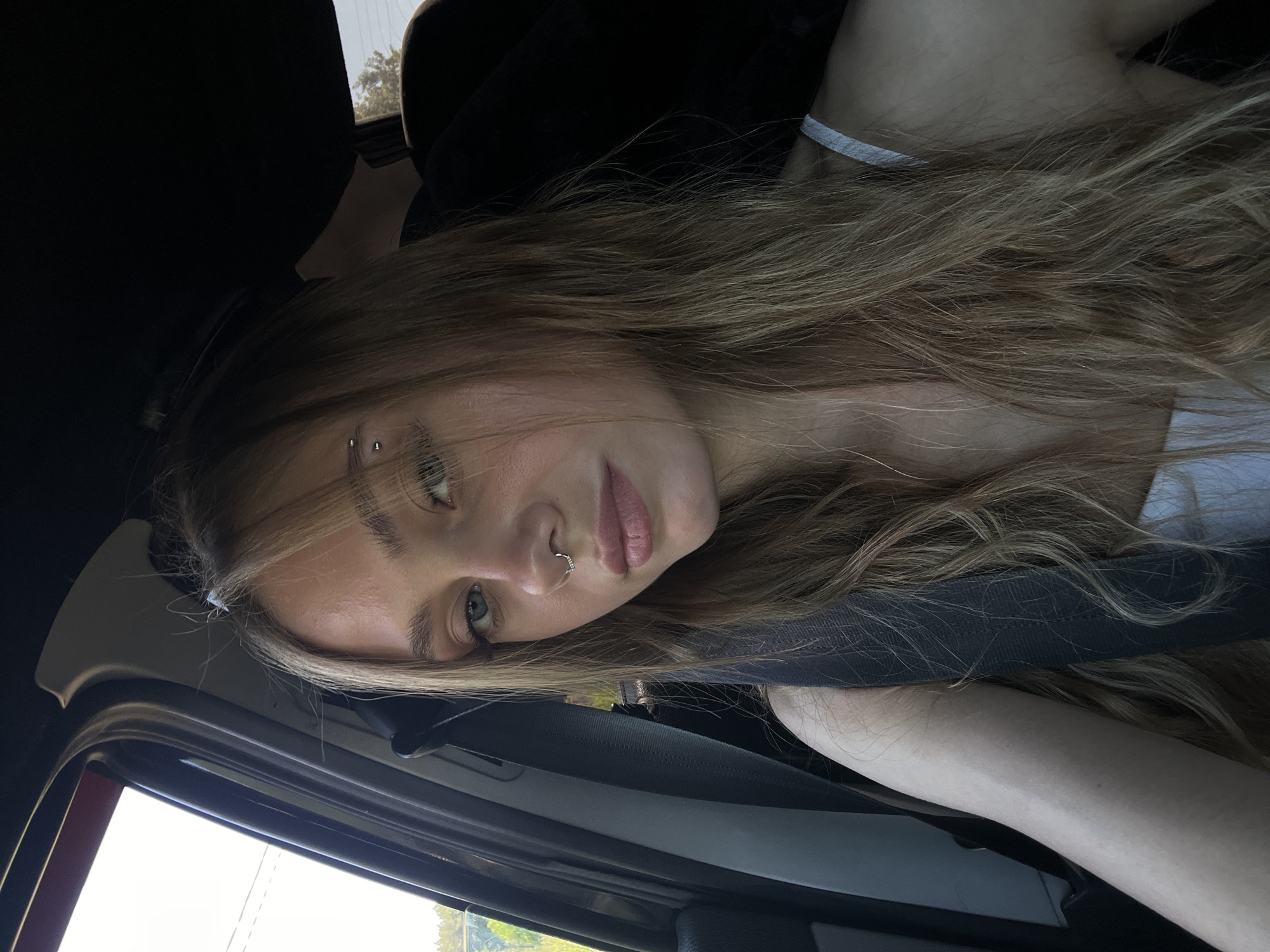 A young woman with blonde hair and blue eyes is sitting in the passenger seat of a car. She is wearing a white tank top and a seatbelt is visible across her chest. She has a nose ring and an eyebrow piercing. Her hair is long and wavy, and it falls around her shoulders. The interior of the car is dark, with the sun shining in through the window on the left. The background outside the window appears to be a natural setting with trees and foliage, suggesting a location on the outskirts of Chaikovsky, Russia.