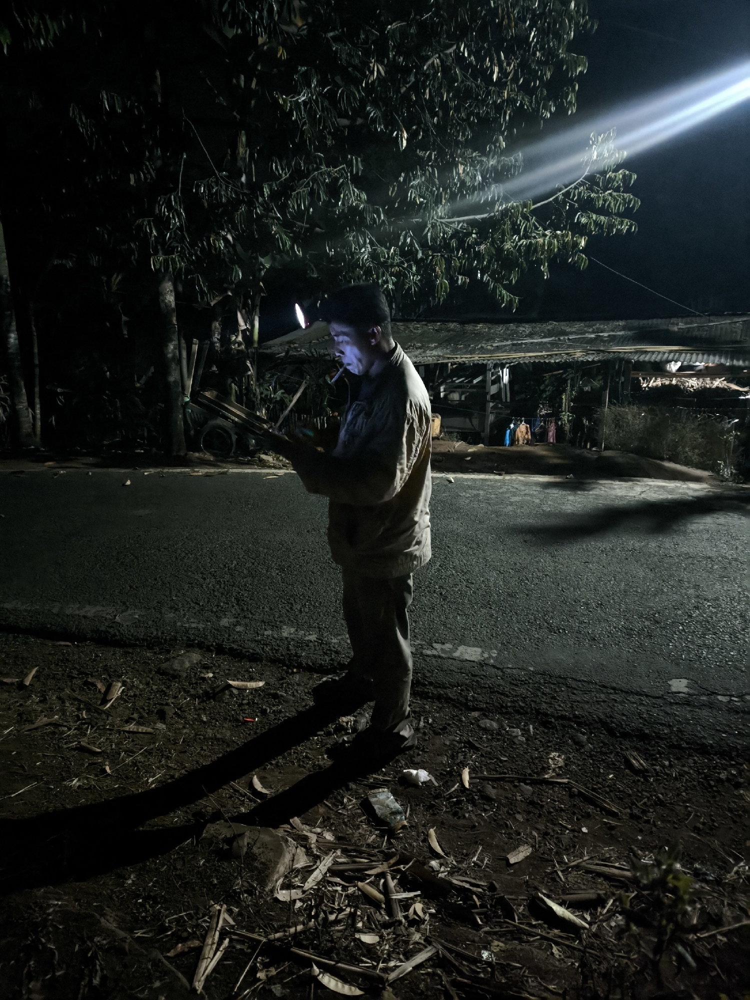 In Bodang, Indonesia, at night, a person stands by the side of a road, illuminated by a headlamp and a distant bright light. The individual is wearing a light-colored jacket and dark pants, holding a mobile phone and what appears to be a cigarette in their mouth. They are looking down at the phone, with their shadow cast on the asphalt road. The surroundings are dark, with trees and vegetation visible. In the background, a simple structure with a corrugated roof and clothes hanging suggests a residential or market area. The ground is covered with dirt, debris, and dry leaves.