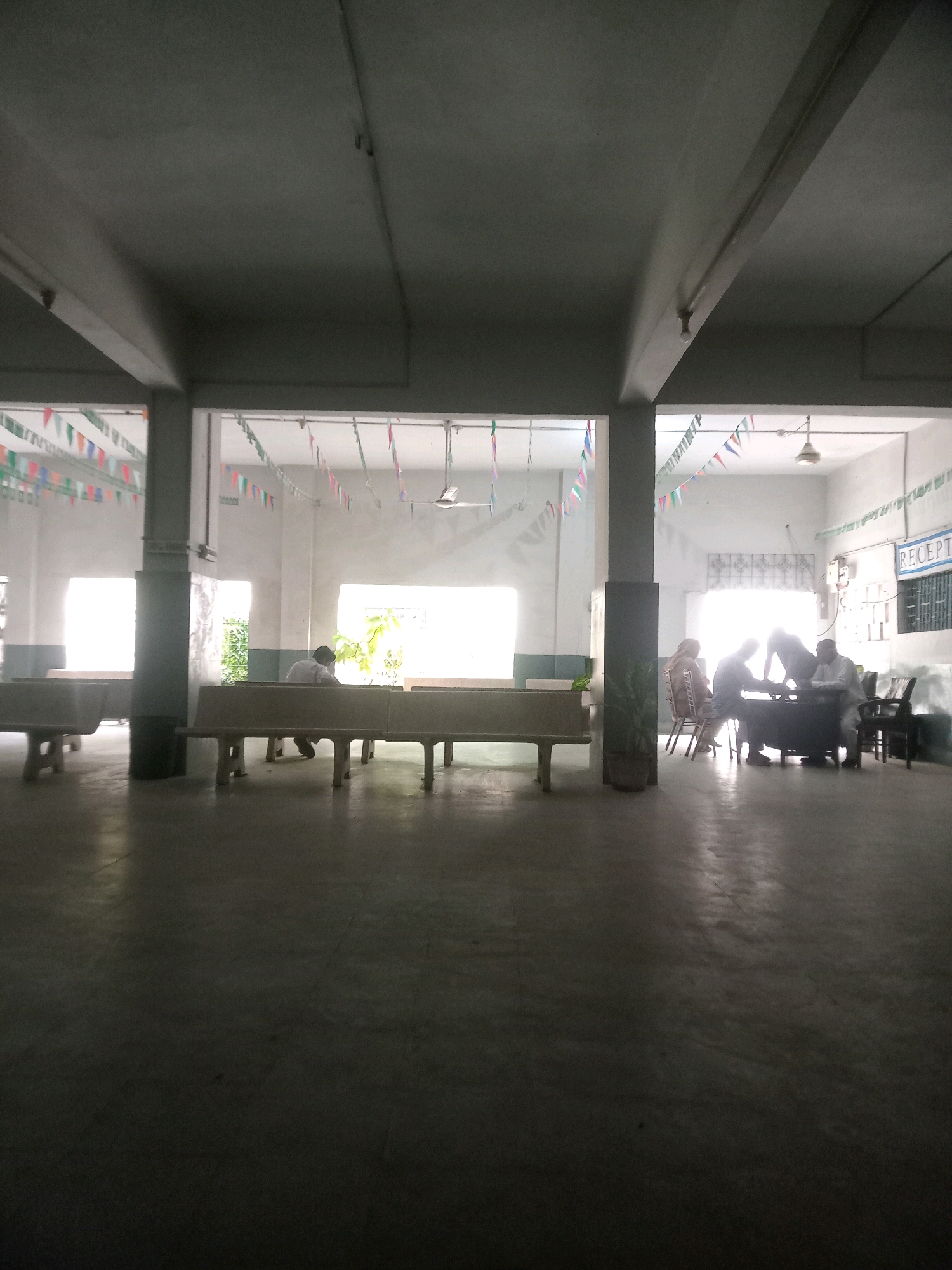 This is a medium shot, eye-level, vertical interior photograph of a waiting area that appears to be in a government office or public institution. The scene is dimly lit, with light filtering in from windows on either side of the frame and from overhead fluorescent lights. Bunting flags are strung across the ceiling in a festive manner.
In the foreground, there are two long, concrete benches with metal legs. The floor is a polished concrete or terrazzo surface.
In the midground, on the left side of the image, a lone individual, presumably male, is seated on one of the benches, facing away from the camera and towards a window. Their posture suggests they are looking down, possibly at a mobile device or reading material.
Further back, in the right midground, a group of four individuals are gathered around a desk. Two appear to be seated at the desk, while two others are standing. One seated individual, a woman in a headscarf, is visible. The individuals are engaged in some form of interaction, possibly administrative or consultative. A sign reading "RECEPT" is visible on the wall behind them, indicating this is likely a reception or administrative area.
The environment appears somewhat dated, with visible structural elements like columns and beams. The overall impression is that of a functional but not particularly modern public space. The setting is consistent with a location in Karachi Division, Pakistan, given the context of typical public buildings in the region and the presence of cultural attire.