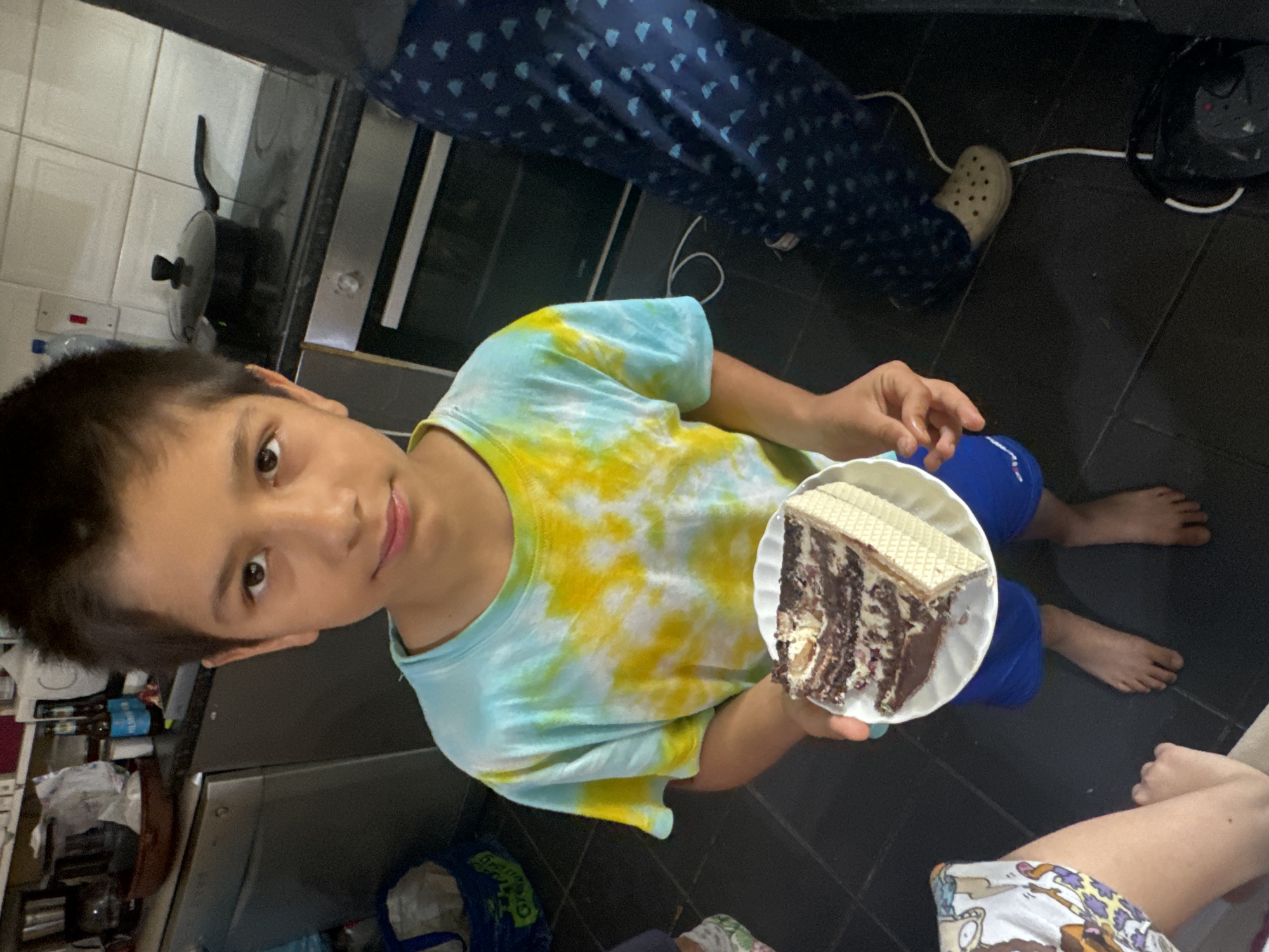 A young boy with dark hair, wearing a tie-dye t-shirt and blue shorts, stands in a kitchen and holds a slice of chocolate cake on a white plate. He is barefoot. To the right of the boy, a person in dark blue patterned pants and beige clogs is partially visible. The kitchen counter behind the boy has a dishwasher and a stove with a black pot on it. The floor is black tile. The scene is indoors. The image does not provide enough information to determine the location in Waterford, Ireland.