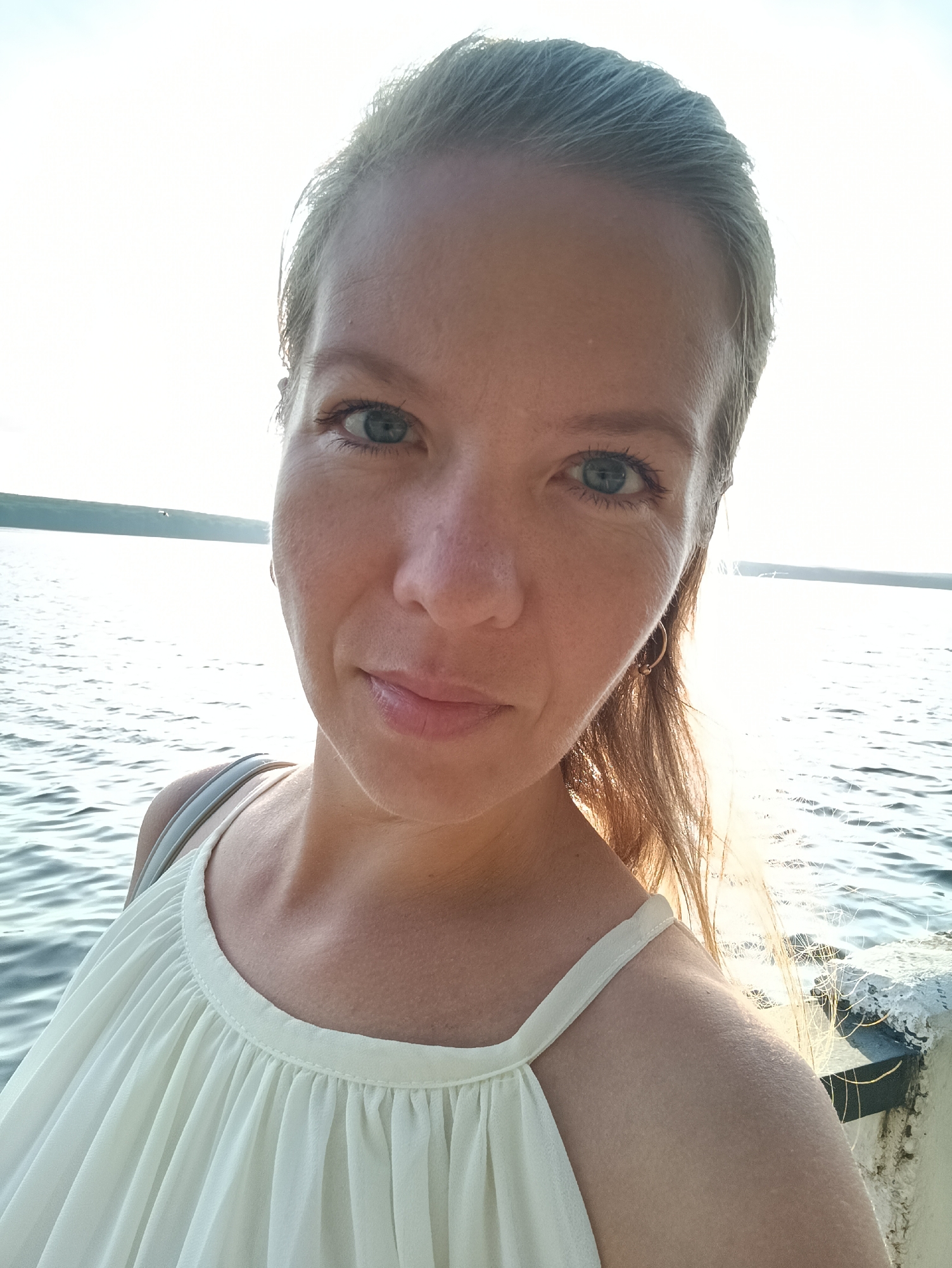A young Caucasian woman with blonde hair pulled back and light blue eyes is taking a selfie. She is wearing a light-colored, gathered-neckline dress. She is positioned in the foreground with a body of water and a hazy horizon in the background. The setting sun casts a warm glow on the scene. The image appears to be taken outdoors, possibly on a pier or a waterfront. The woman is smiling slightly and looking at the camera.