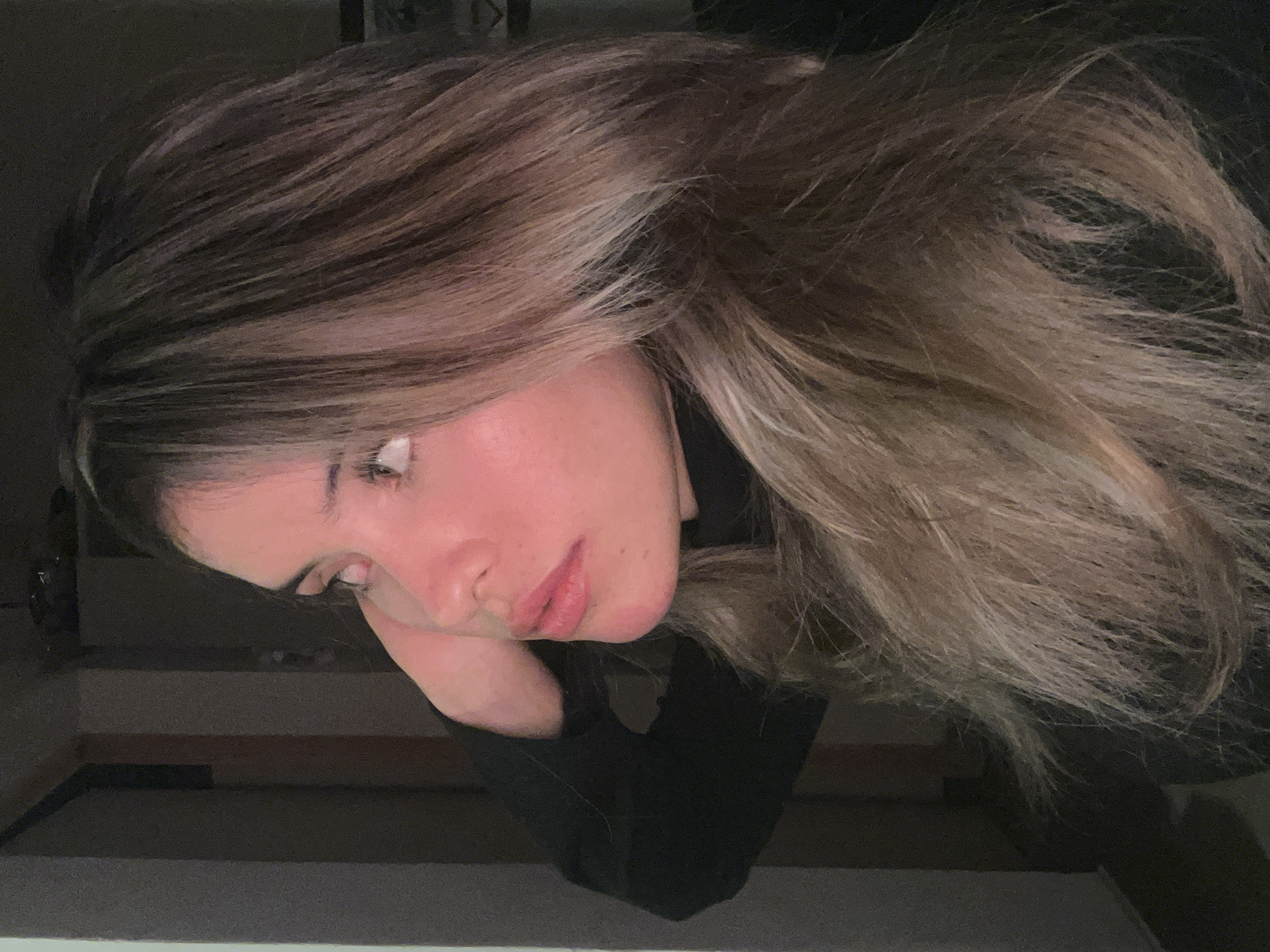A young woman with blonde hair is seen in a profile view, looking up and to the left. Her right hand is placed behind her head, and she is wearing a black turtleneck. The background appears to be indoors, with dimly lit walls and some shadow. There is no indication of the city of Sumalao, Argentina, in the image or its context.
