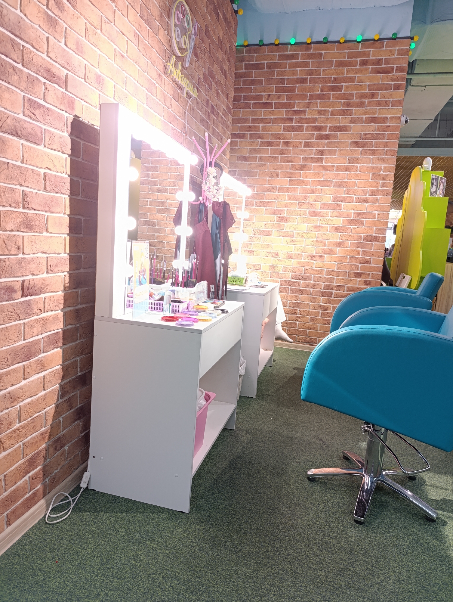 The image shows a children's makeup studio in an indoor setting, likely within an entertainment or recreational facility. The studio features a white vanity table with a light-up mirror, stocked with various makeup items and brushes. Behind the vanity, a mannequin is dressed in a burgundy garment, possibly for dress-up or role-play purposes.
To the right of the vanity, two turquoise chairs are positioned, indicating seating for patrons receiving makeup services. The walls are constructed of brick, and decorations include illuminated spheres.
The flooring is carpeted in a dark green. An electrical cord is visible near the base of the vanity table, suggesting it is connected to a power source for the lights.