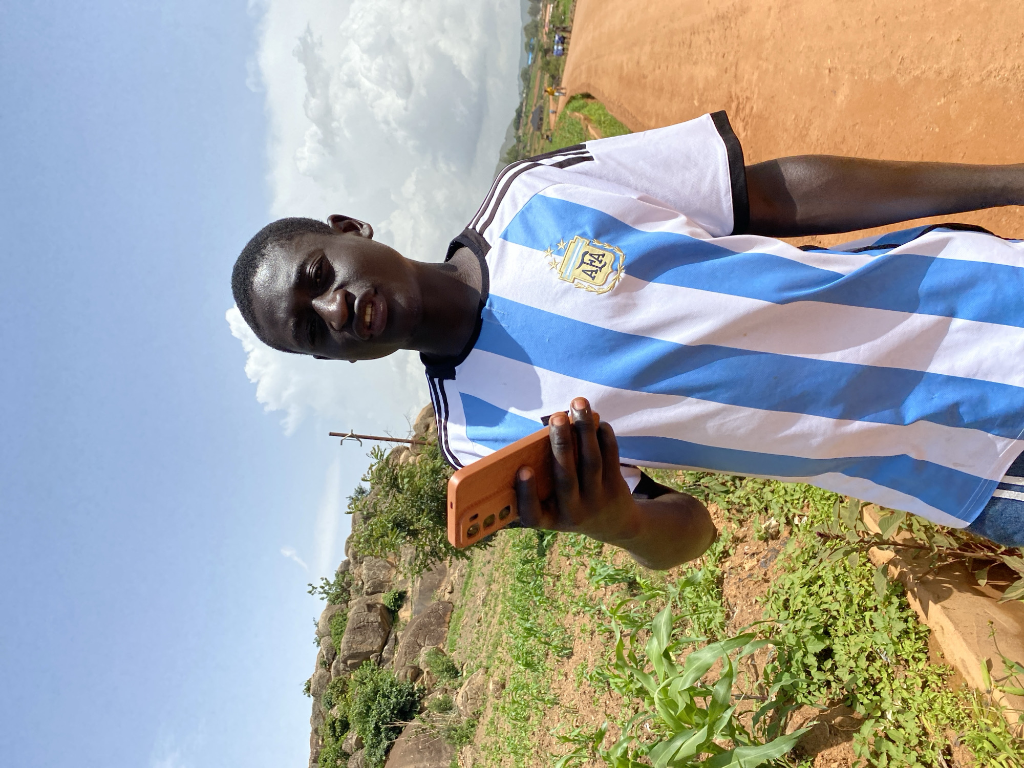 A young Black male is pictured outdoors in Bukuru, Nigeria, holding a smartphone. He is wearing a white and light blue striped t-shirt with black stripes on the shoulders and the AFA logo on the chest, likely an Argentina national team jersey. The man has short dark hair and is looking towards the camera with a neutral expression.
The setting appears to be a rural or semi-rural area. The ground is a mix of reddish-brown soil and green vegetation, with some rocks and larger plants visible. In the background, there is a dirt road or path with a few people walking on it, and some buildings and trees further in the distance. The sky is blue with scattered white clouds. The lighting suggests it is daytime, possibly late morning or early afternoon, with bright sunlight. The overall mood is casual and relaxed. No visible text is present beyond the logo on the shirt.