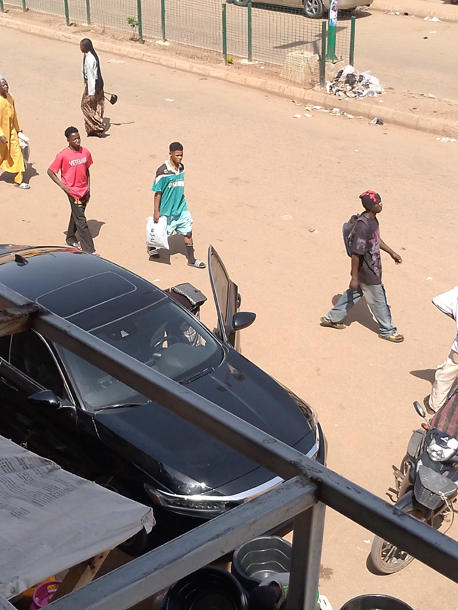 The image shows a street scene in Bukuru, Nigeria. The ground is a light brown color. Several people are walking on the street. A man in a yellow robe, a man in a red shirt, a man in a teal shirt, a man in a black shirt with a backpack, and a man in a white shirt. A black car with an open driver-side door is parked on the side of the street. A motorcycle is parked next to the car. A pile of trash is on the side of the road. A metal fence is visible in the background. The image appears to be taken from an elevated position. Some metal framing and buckets are visible in the foreground.