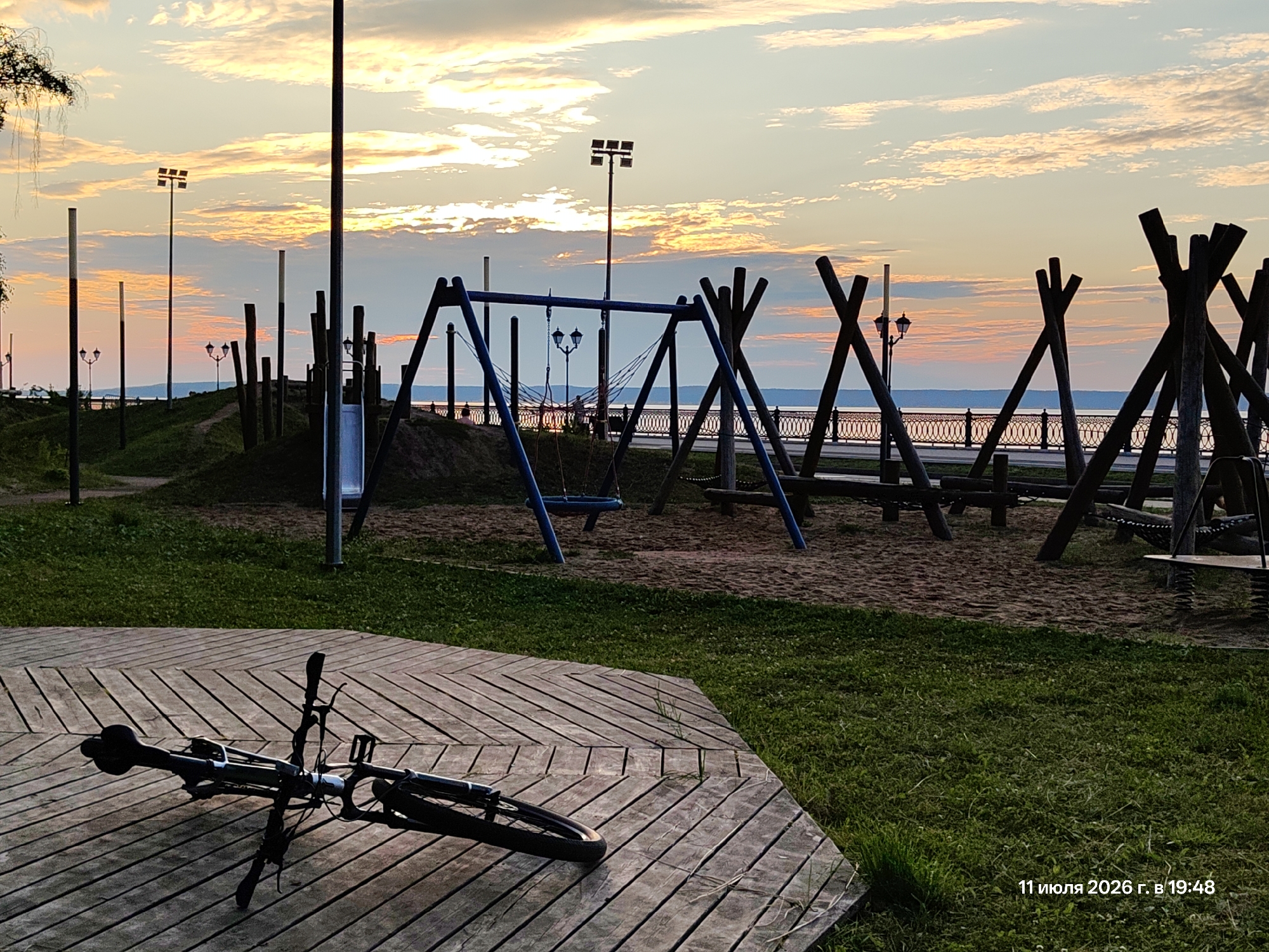 The image captures an outdoor scene at dusk, depicting a playground and a waterfront promenade. In the foreground, a bicycle lies on its side on a wooden deck. The playground features swings and climbing structures made of wood and metal, set against a sandy base. Beyond the playground, a body of water is visible, with a decorative railing along the promenade. The sky is illuminated by a sunset, with colorful clouds and the silhouettes of lampposts and trees. The overall atmosphere suggests a peaceful evening in a public park. The timestamp "11 июля 2026 г. в 19:48" is visible in the bottom right corner, indicating the date and time the photograph was taken.