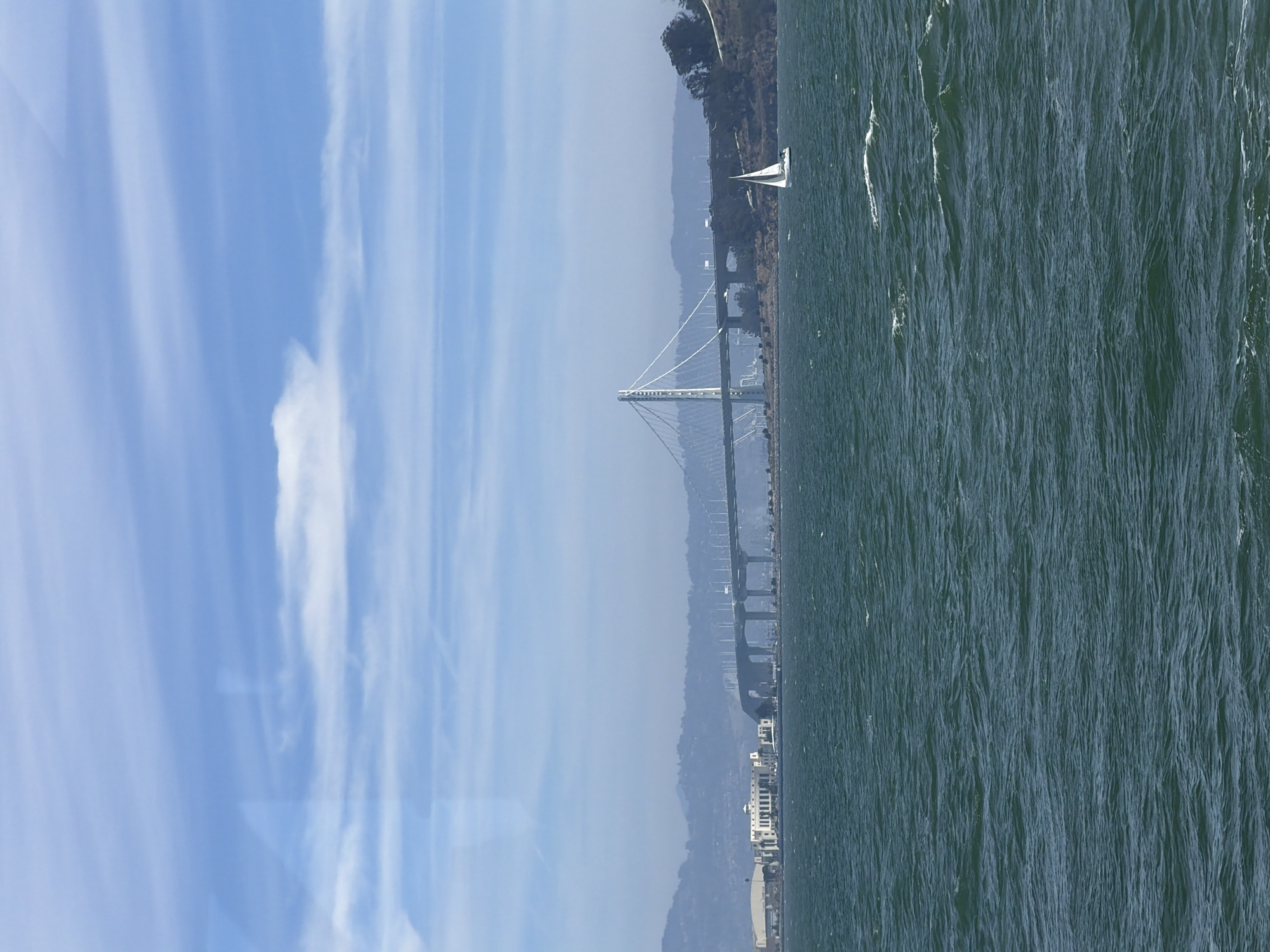 This is a landscape photograph taken from the water, likely the San Francisco Bay, looking towards the city and its landmarks. In the foreground, the choppy, dark teal water of the bay dominates the lower half of the image, with whitecaps suggesting a breezy day.
A white sailboat with its sail up is visible in the middle ground, moving from right to left. Beyond the sailboat, the Bay Bridge is prominently featured, stretching across the water. The bridge has a distinctive cable-stayed design with a tall white tower. Cars can be seen on the bridge, indicating active traffic.
On the shoreline to the left of the bridge, several modern buildings with flat roofs and white facades are visible, possibly representing a corporate campus or a part of the city's infrastructure. Further in the distance, rolling hills form a hazy backdrop, suggesting the landscape surrounding the bay.
The sky above is a light blue, with scattered white, wispy clouds. The overall impression is a bright, possibly slightly overcast day with a moderate amount of wind. The scene captures a classic view of the San Francisco Bay, highlighting its iconic bridge and maritime activity.