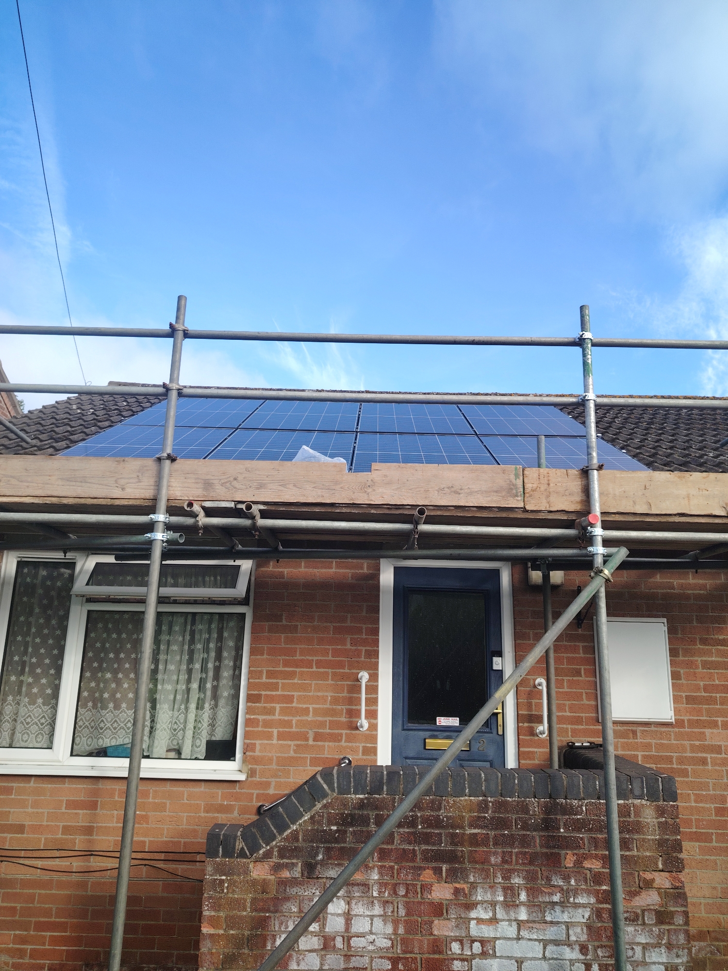 The image displays the front facade of a two-story red brick residential house under a bright blue sky with scattered cirrus clouds. A thin, dark overhead power line diagonally crosses the upper left quadrant of the sky.
The roof, covered in dark tiles, features an array of twelve dark blue solar panels centrally mounted. A white protective sheet is visible beneath the panels in a small section on the lower right of the array, indicating recent installation or ongoing work.
Scaffolding is erected across the entire width of the house. Two main vertical metal poles are prominent in the foreground, supporting horizontal poles and a wooden plank platform at approximately the eave level, positioned just below the solar panels. Various metal clamps and connectors secure the scaffolding elements. A red cap is visible on the end of a horizontal scaffolding pole on the right side.
The house itself is constructed from red brick laid in a stretcher bond pattern. The lower brick courses show patches of white efflorescence and general weathering. On the left side of the ground floor, there are two white UPVC casement windows, each containing two panes. Sheer curtains with a star or snowflake pattern are visible through the windows. The top-right pane of the left window is slightly ajar.
A dark blue front door, framed by white trim, is centrally located on the ground floor. It features a gold-colored letterbox and handle. A circular doorbell or camera device is mounted to the left of the door, and a white sticker on the door reads "NO JUNK MAIL". The house number "2" is partially visible below the handle. Two white grab handles are affixed to the brick wall, one to the left and one to the right of the front door. To the right of the door, a white rectangular utility box is mounted on the brickwork.
A low brick wall, capped with dark bricks, forms a raised platform or steps leading to the front door. A dark, possibly plastic, storage object or bin rests on this coping to the right of the door. Angled scaffolding support bars extend into the foreground on the right.
The scene is consistent with a residential property in Huntingdonshire, United Kingdom, undergoing external works such as solar panel installation.