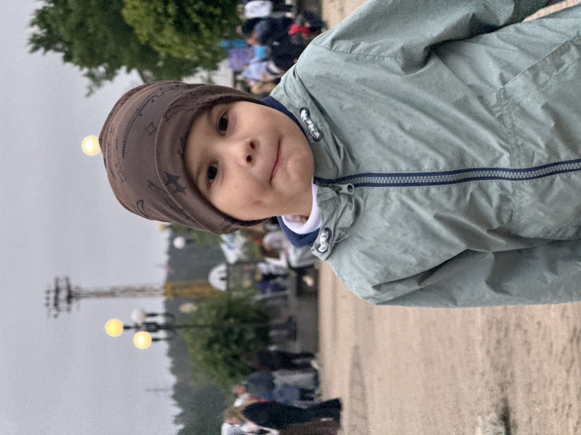 A young child wearing a brown patterned beanie and a light blue jacket is the main subject of this medium shot, taken outdoors. The child is looking slightly towards the camera with a small smile. They have dimples on their cheeks. The jacket has a dark blue zipper and trim. In the background, slightly out of focus, are other people and trees. Several lampposts with glowing yellow lights are also visible, suggesting it might be evening or a cloudy day. The overall setting appears to be a park or public space. The image is oriented vertically, with the child's head towards the top left. There is no visible text.