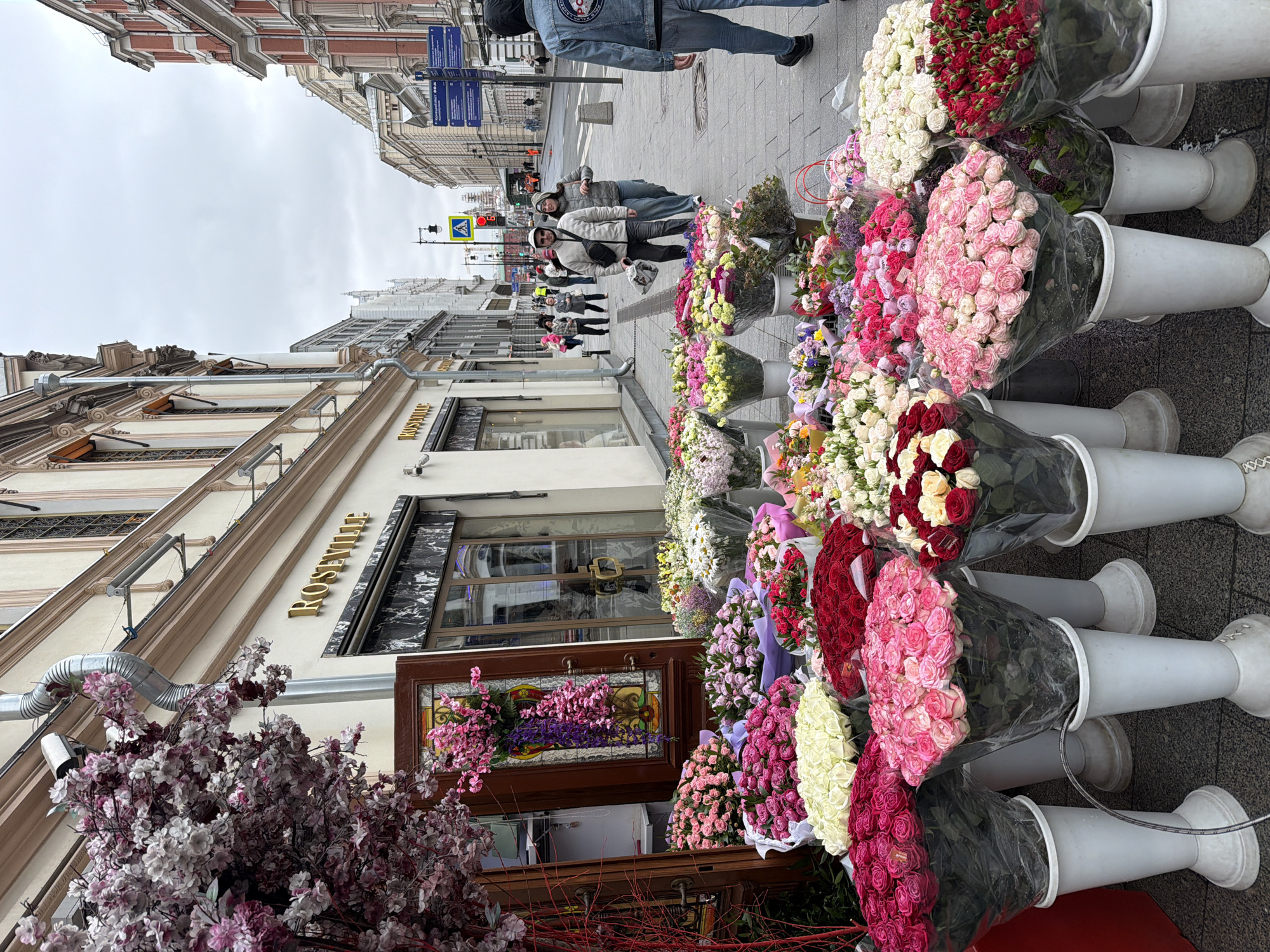 A street-level view shows a flower shop's display in Moscow, Russia. Bundles of roses in white, red, pink, and mixed colors are arranged in white vases along the sidewalk. A building with the gold-lettered sign "ROSEVILLE" stands beside the display. A wooden-framed display is beside the flower shop with flowers inside. On the sidewalk, pedestrians walk past the display. The sky is overcast, and other buildings are visible in the background along the street.