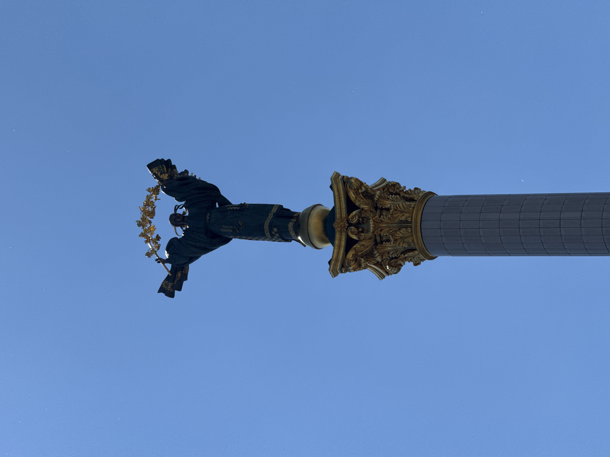 The image captures the Monument of Independence, located on Maidan Nezalezhnosti in Kyiv, Ukraine. The monument features a tall, dark grey column adorned with ornate golden detailing at its top. Perched atop the column is a bronze statue of a woman in a flowing robe, holding a golden wreath aloft in her outstretched arms. The statue and column are set against a clear, bright blue sky, with no other elements visible in the frame. The perspective is from below, looking up at the monument.