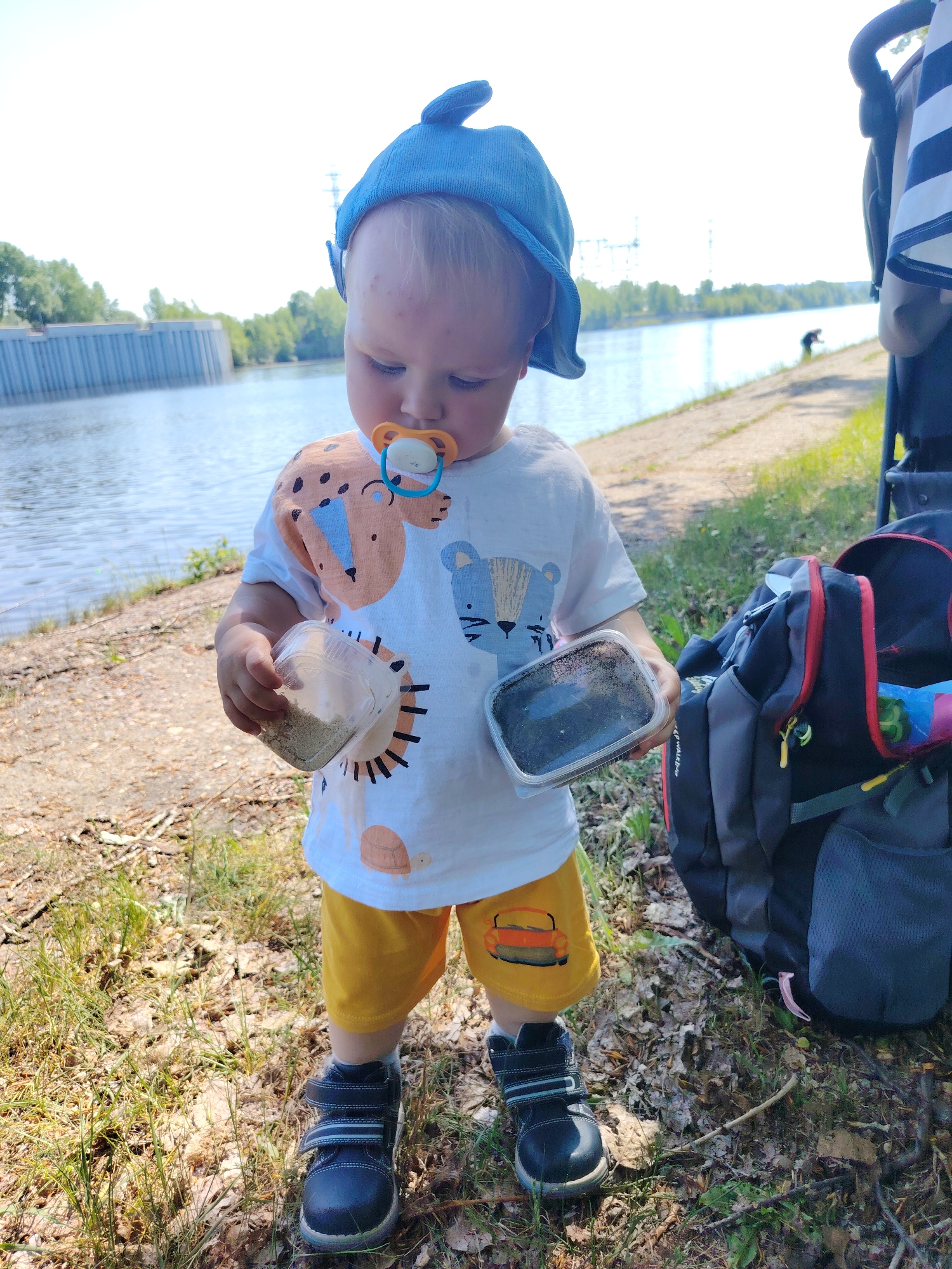 The image shows a toddler standing on a grassy and leafy ground near a body of water. The toddler wears a blue shark-shaped hat, a white t-shirt with animal prints, yellow shorts with a car design, and dark blue shoes with Velcro straps. The toddler holds a clear plastic container with light-colored material in the right hand and a black container in the left. A pacifier is in the toddler's mouth. A gray and black backpack is to the right of the toddler. In the background, there's a wide body of water, a concrete structure, trees, and what appears to be power lines. The location is Chaikovsky, Russia.