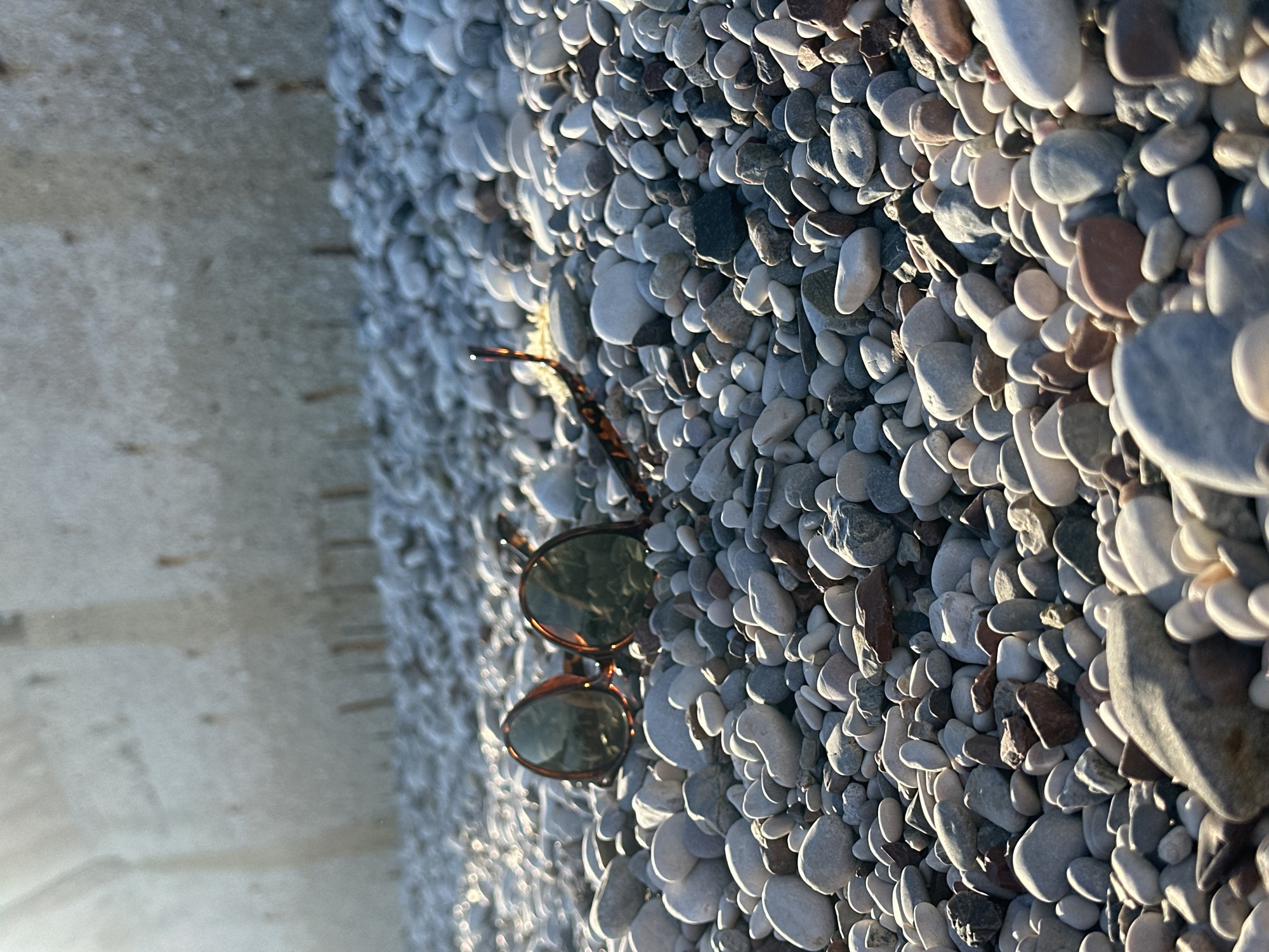 The image shows a close-up of a beach scene in Budva, Montenegro. The foreground is dominated by smooth, oval-shaped pebbles and stones of varying sizes, primarily grey and white with some brown and darker shades. Sunlight hits the pebbles, creating shadows and highlights. Sunglasses lie on the pebbles; they have a tortoiseshell frame and dark lenses. To the left of the pebble beach is a concrete surface. The scene appears to be outdoors on a sunny day.